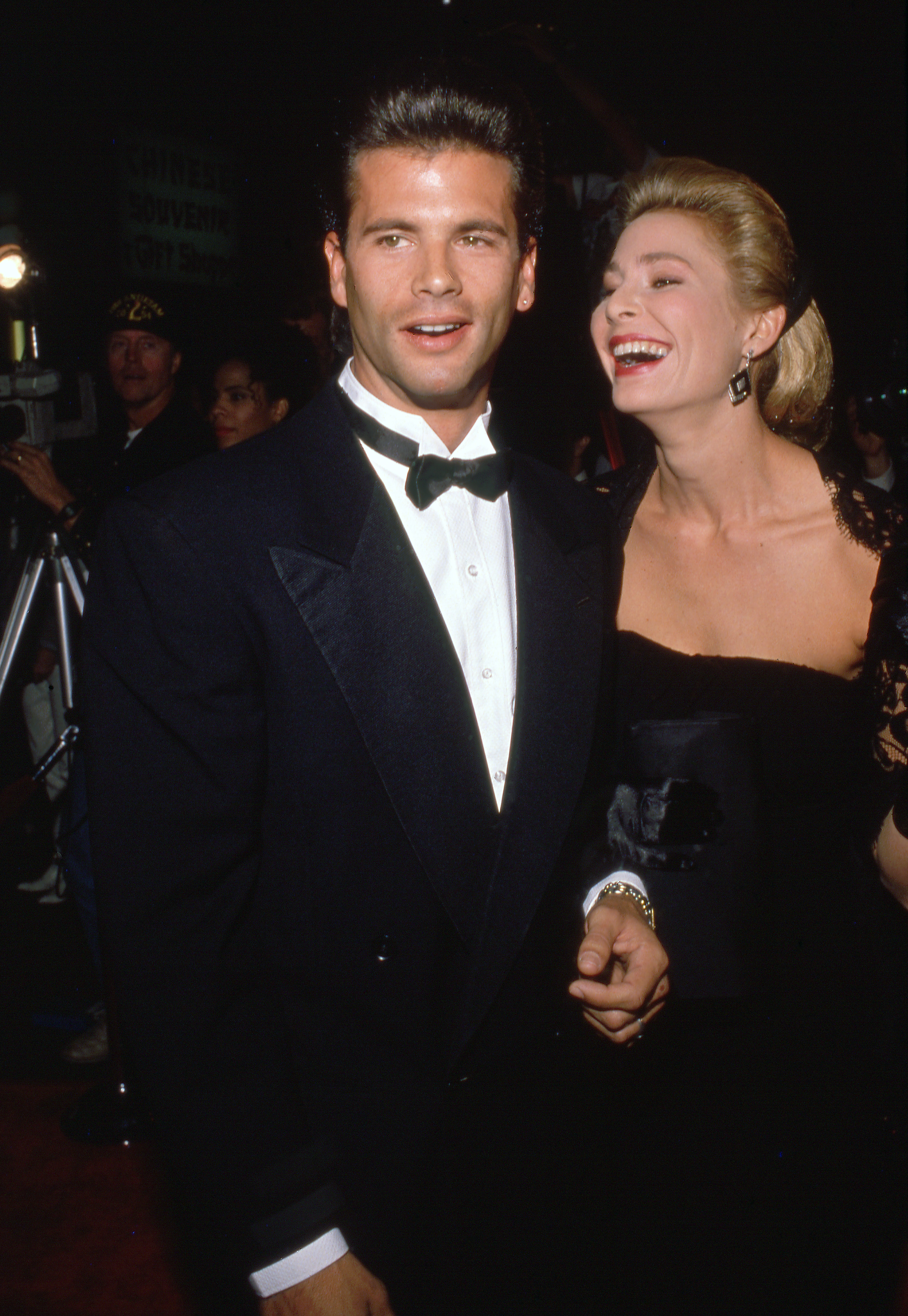 Lorenzo Lamas and his then-wife, actress Kathleen Kinmont, share a joyful red-carpet moment during a 1990s evening event. Lamas looks polished in a classic black tuxedo, white pleated shirt, and black bow tie, while Kinmont laughs beside him in an elegant black dress, swept-back blonde hair, and statement earrings. | Source: Getty Images