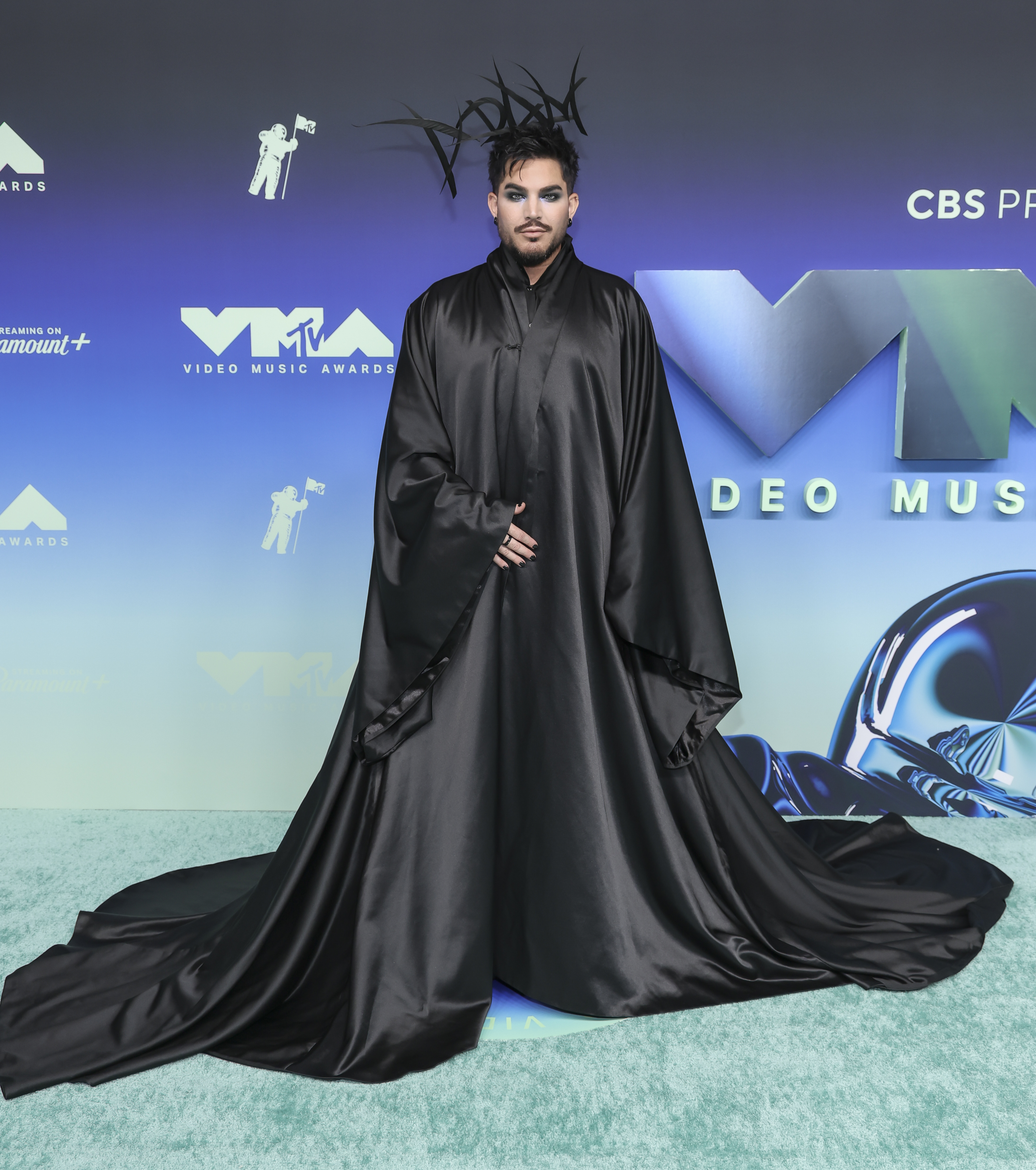 Lambert posed beneath a dramatic sweep of black fabric as his floor-length cape pooled across the carpet. Long, pointed spikes in his hair extended the silhouette upward, creating a sharp contrast with the cape's broad shape below. | Source: Getty Images