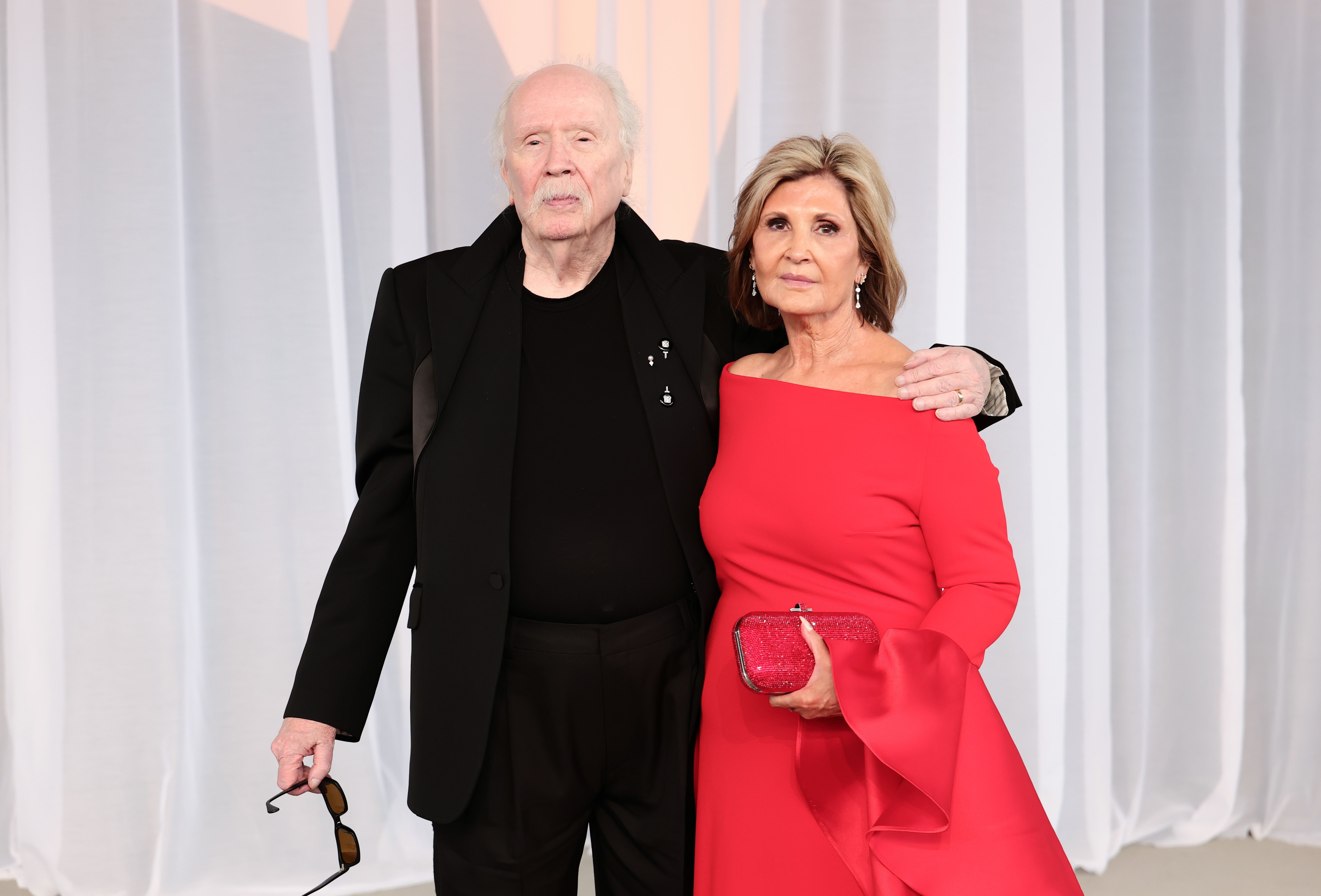 John Carpenter and his wife, producer Sandy King, pose together in coordinated style. King stands out in a vibrant cherry red off-the-shoulder gown featuring dramatic bell sleeves and paired with a glittering ruby clutch, while Carpenter opts for an all-black tailored suit with sharp lapels and a matching dark crewneck, holding his sunglasses at his side as he celebrates receiving the Luminary Award.