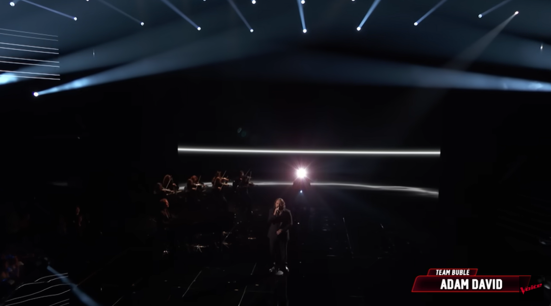 A wide shot of the stage shows Adam David singing under minimal lighting, with the orchestra arranged behind him in silhouette | Source: YouTube/nbcthevoice