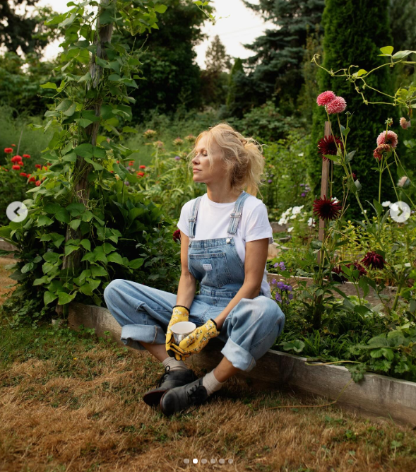 Pamela Anderson pauses for a moment of quiet reflection in her garden, sitting cross-legged on a raised bed in denim overalls, floral gloves, and lace-up boots while cradling a mug in the afternoon sun. Surrounded by tall blooming dahlias, lush greenery, and runner beans, she shared the tranquil scene alongside a note celebrating her food series "Pamela's Cooking with Love" as "fun, fun, fun". | Source: Instagram/pamelaanderson