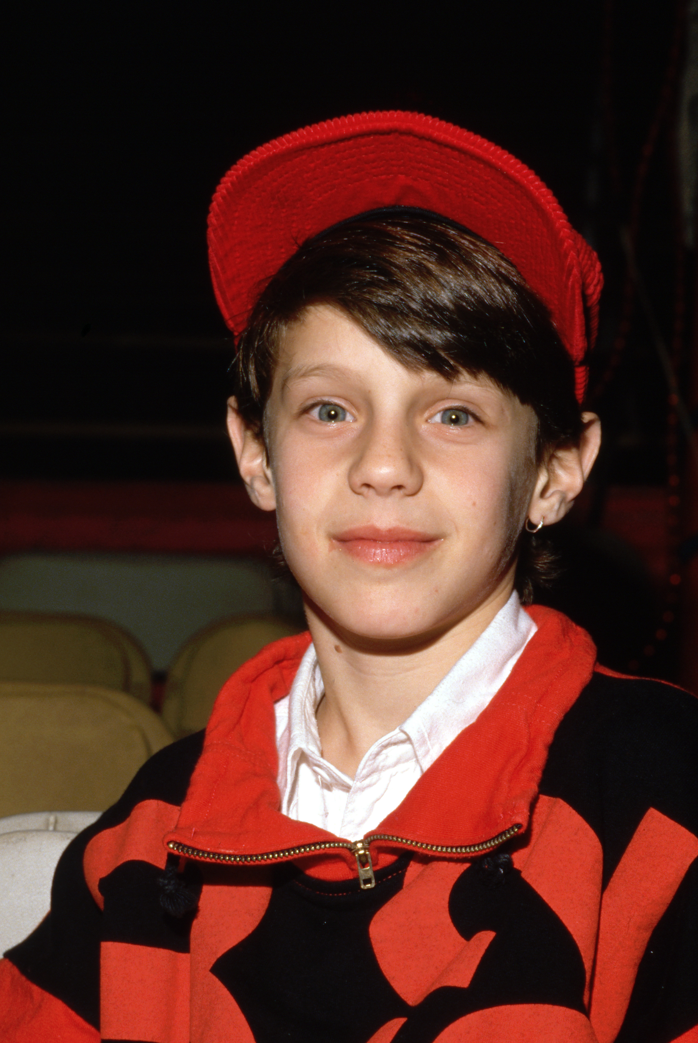 Omri Katz shows off a playful preteen look with side-swept dark bangs peeking out from beneath a bright red cap, a small hoop earring in his left ear, and a white collared shirt layered under a bold red-and-black zip sweatshirt. The portrait captures the young actor attending an event in February 1989 during his long-running role as John Ross Ewing III on the television series "Dallas".