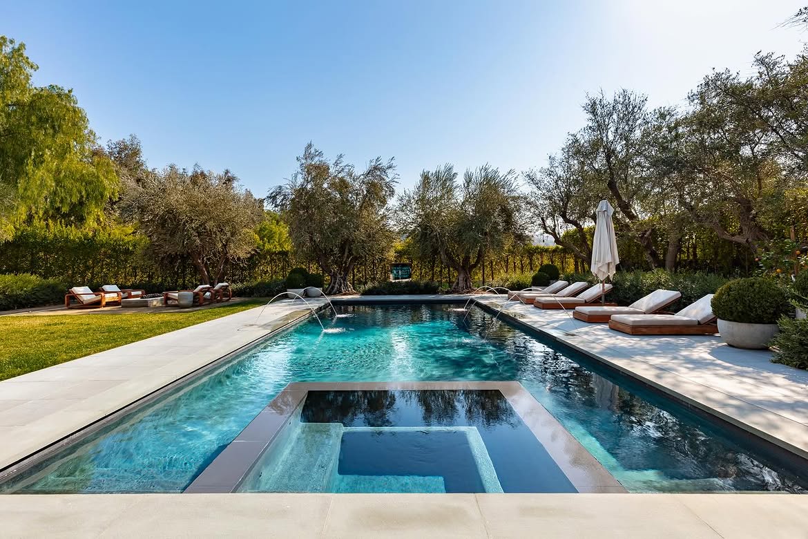 A raised spa anchors one end of the rectangular pool, while curved water features reach across the surface from both sides. Lounge chairs, lawn and dense greenery create several distinct areas around the water. | Source: Instagram/theshevins