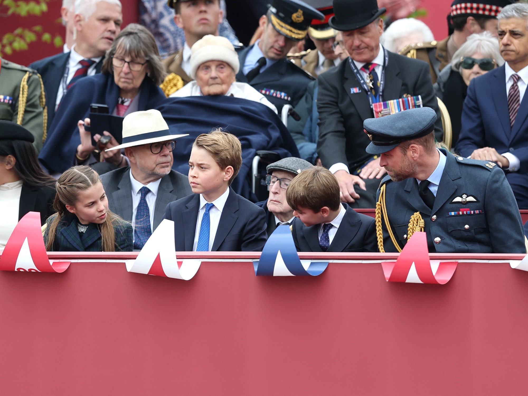 During the VE Day commemoration, Charlotte leans forward in her seat while George looks at her. Louis turns to watch his older siblings while William watches over them. The setting is filled with veterans and officials, emphasizing the solemnity of the occasion.