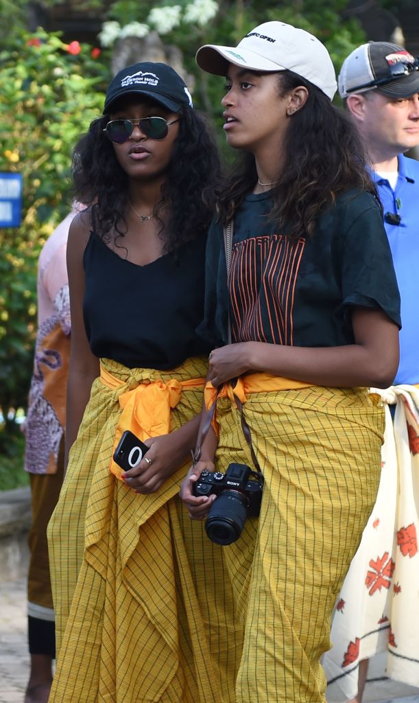 While visiting Bali’s Tirtha Empul temple, Sasha — wearing a black tank, sunglasses, and sarong — holds her phone beside Malia, who carries a camera and looks ahead attentively in a white cap and striped tee tied with a yellow sash.