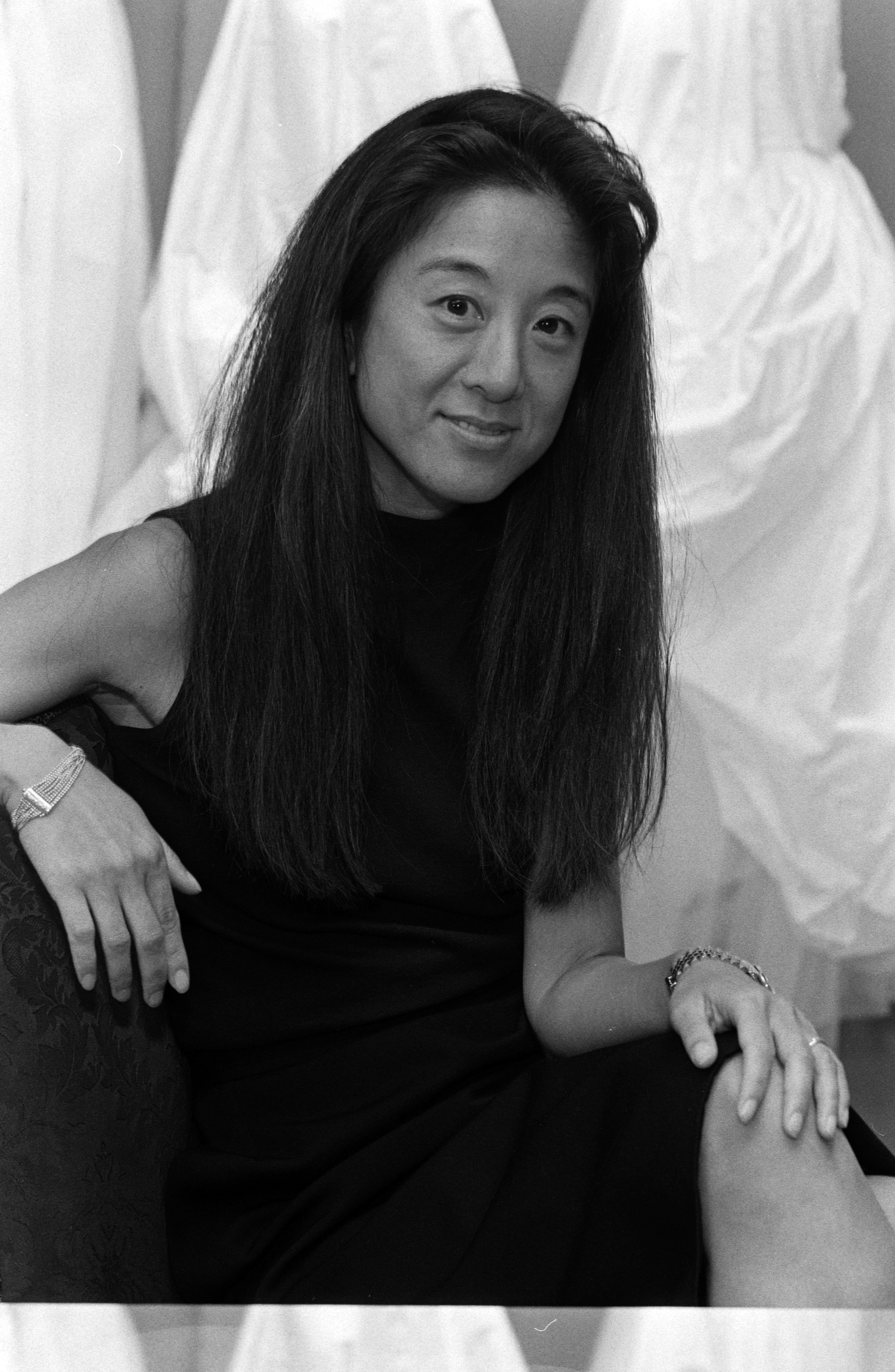 Fashion designer Vera Wang sat for a black-and-white portrait during an interview discussing her foray into the bridal industry and the launch of the Vera Wang Bridal House on August 22, 1990. Seated before draped wedding gowns, Wang smiled softly in a sleeveless dark dress, accessorized with bracelets on both wrists and her signature long dark hair worn straight past her shoulders. | Source: Getty Images