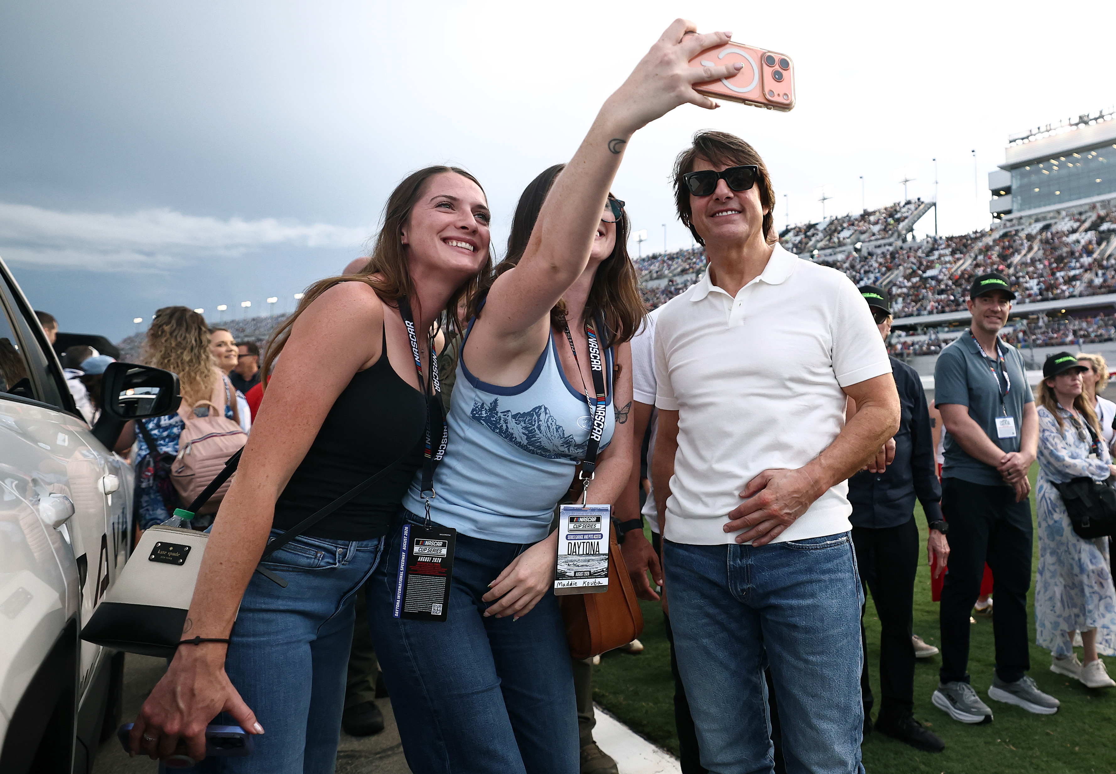 Cruise poses with racegoers for a trackside selfie before the Coke Zero Sugar 400. Wearing sunglasses and a white polo, he stands beside the group with packed grandstands and an overcast sky behind them. | Source: Getty Images