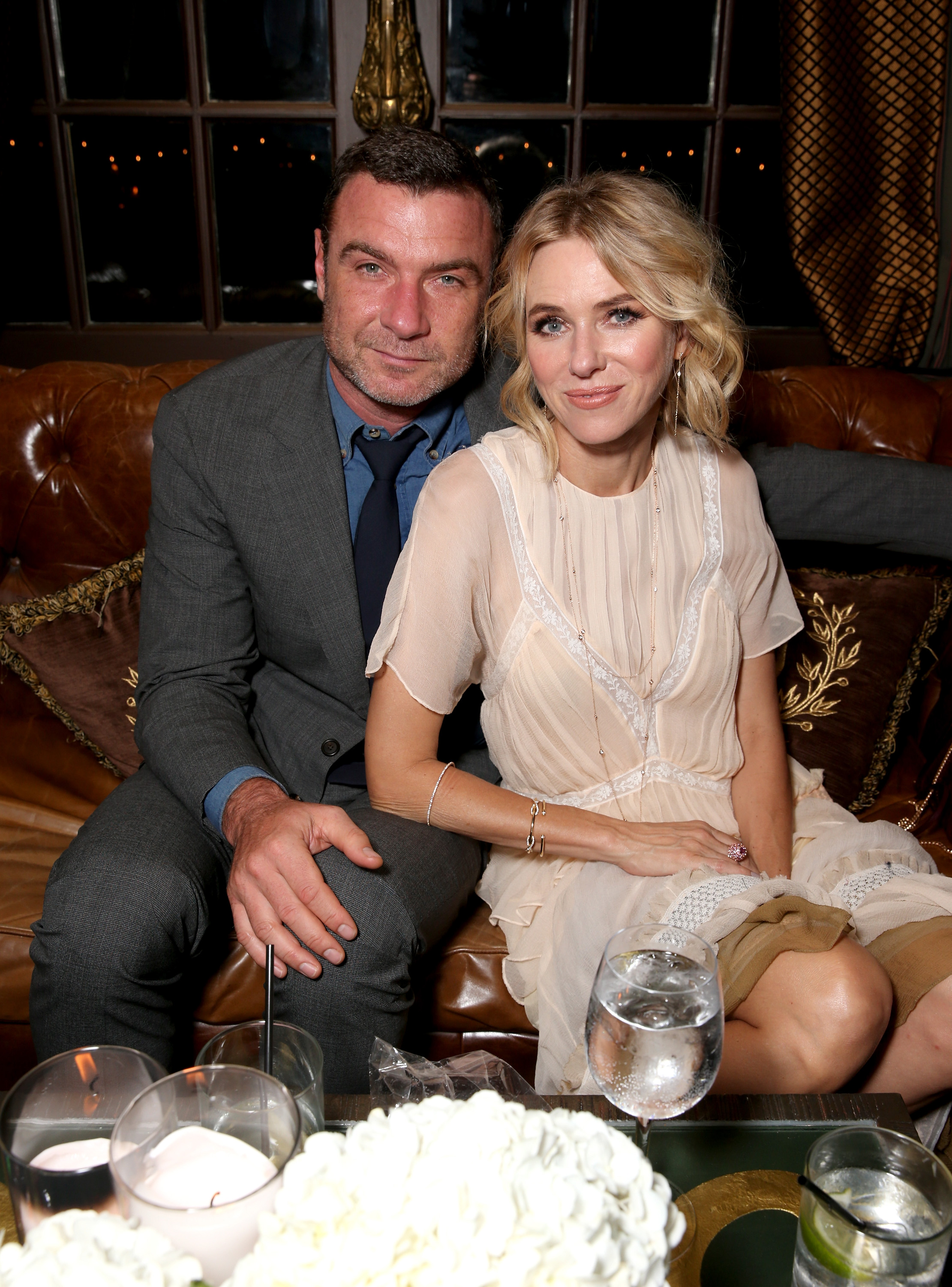Naomi Watts and Liev Schreiber sit closely together at an intimate evening gathering. Watts wears a soft, elegant cream dress while Schreiber appears in a tailored gray suit. The pair lean toward each other on a leather sofa, creating a relaxed and cozy moment.