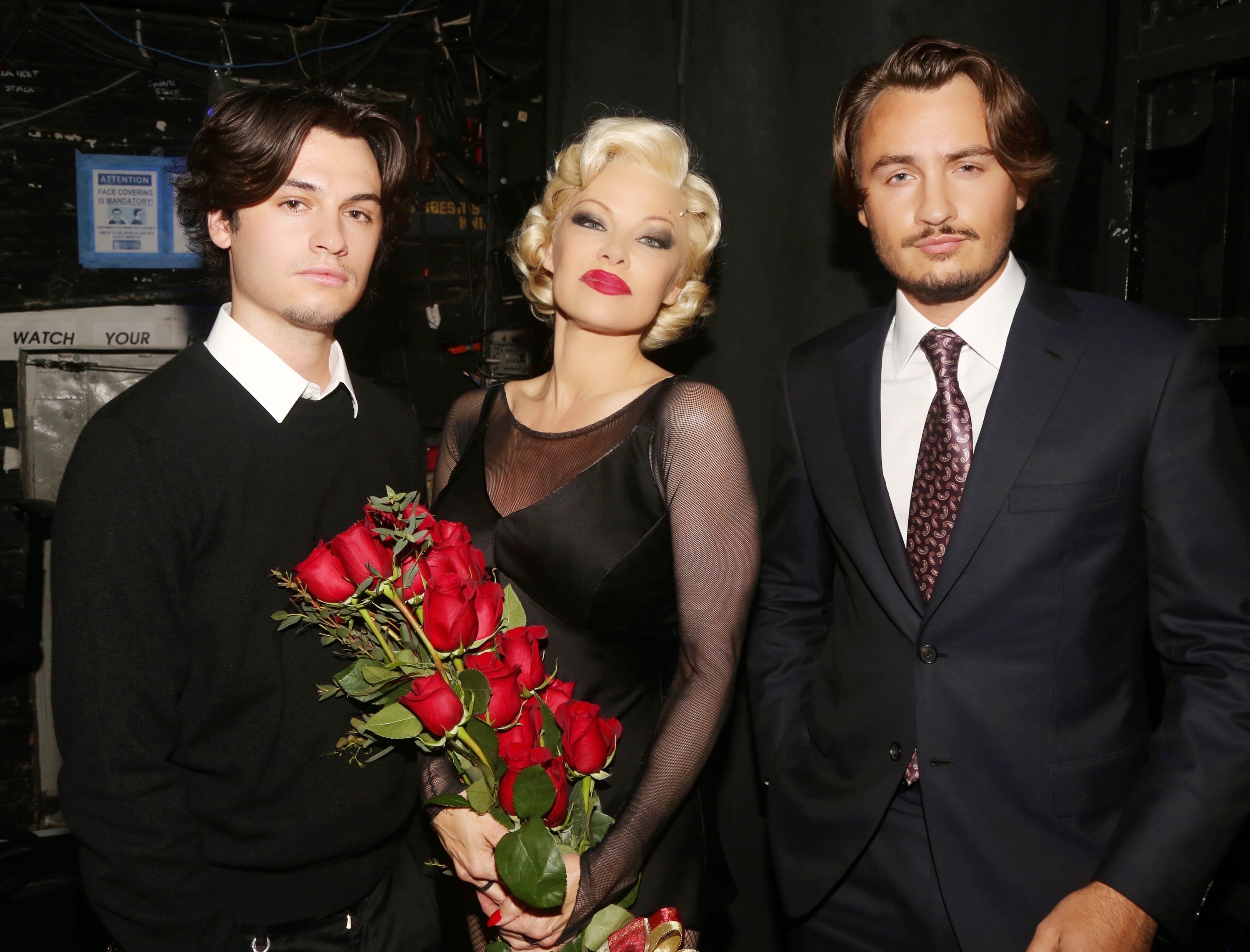 Dylan, Pamela, and Brandon pose backstage on April 12, 2022, during the opening night of her Broadway debut as Roxie Hart in the musical "Chicago" at the Ambassador Theatre in New York City. Pamela holds a bouquet of red roses while dressed in a sheer black costume, flanked by her sons in formal black attire.