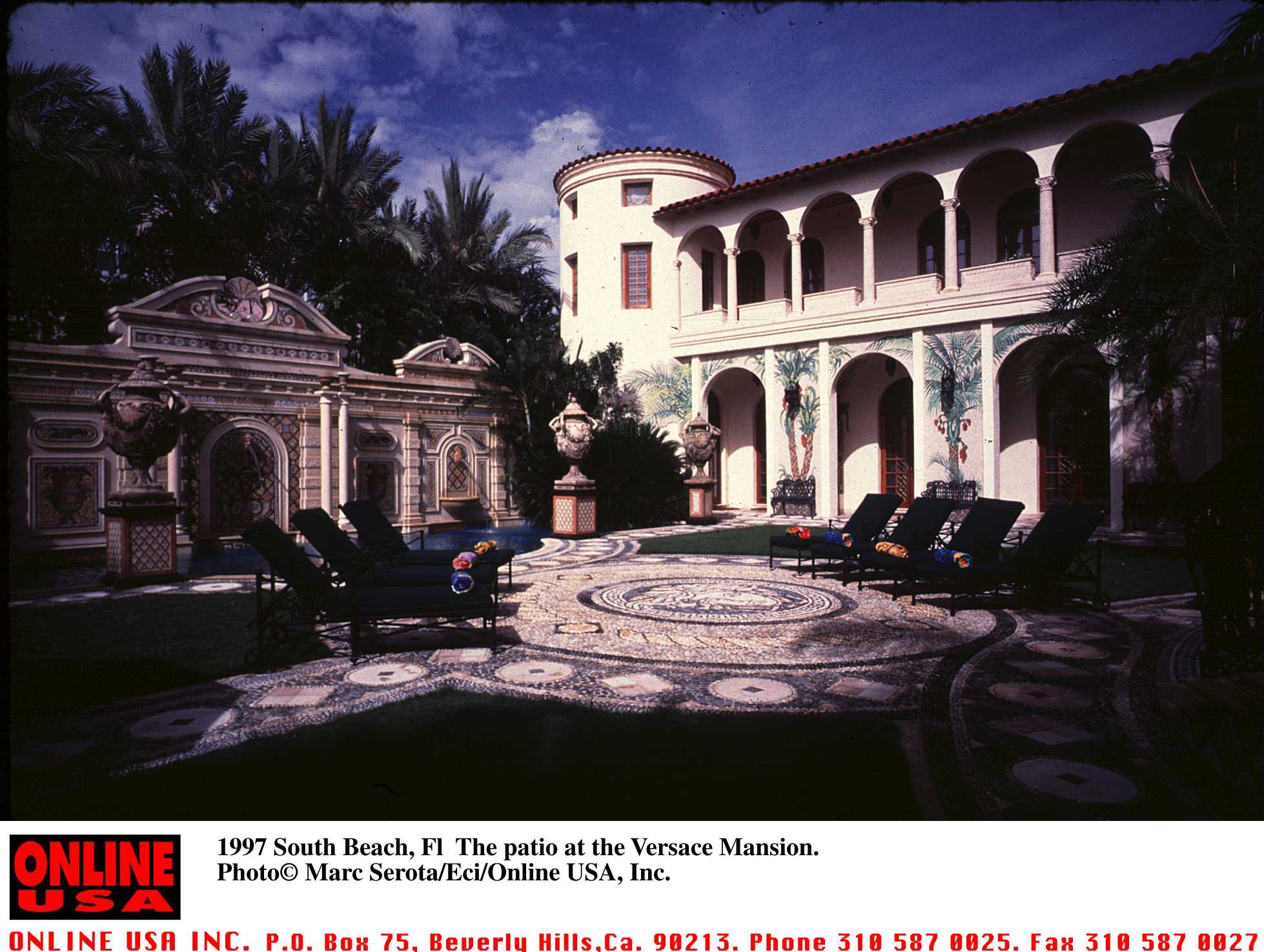 A scenic view captured the lavish courtyard and patio area at the Versace Mansion (The Villa Casa Casuarina) in South Beach, Florida, in 1997. The opulent space features intricate mosaic tile circular paving, dark lounge chairs arranged poolside, ornate classical statues and urns, and a grand Mediterranean-style facade detailed with arched loggias, palm tree wall murals, and a circular tower under a sunny sky. | Source: Getty Images