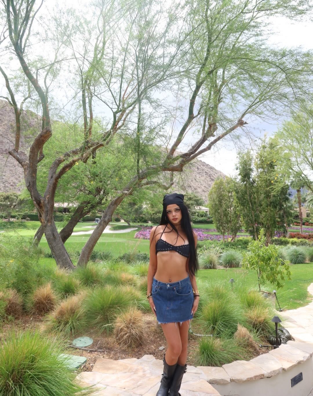 Harlow Winter Kate Madden stands outdoors in a black crop top, denim mini skirt, boots, and a dark headscarf. Framed by trees and desert landscaping, Kate poses confidently. | Source: Instagram/katemaddennn