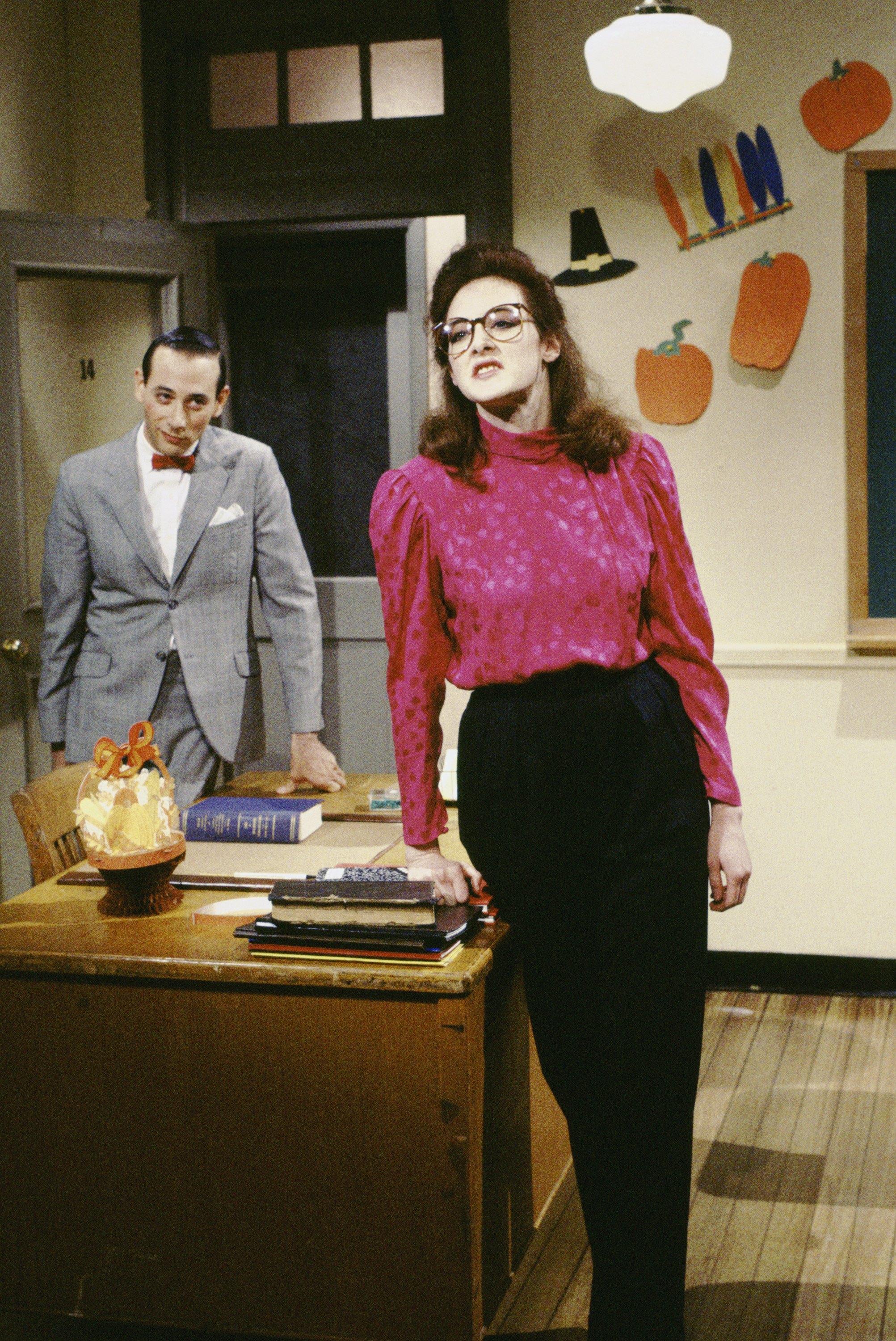Joan Cusack commands the room in a vibrant magenta blouse and oversized glasses while leaning against a desk. Behind her, a suited Pee-wee Herman watches as she delivers a confident, theatrical pose.