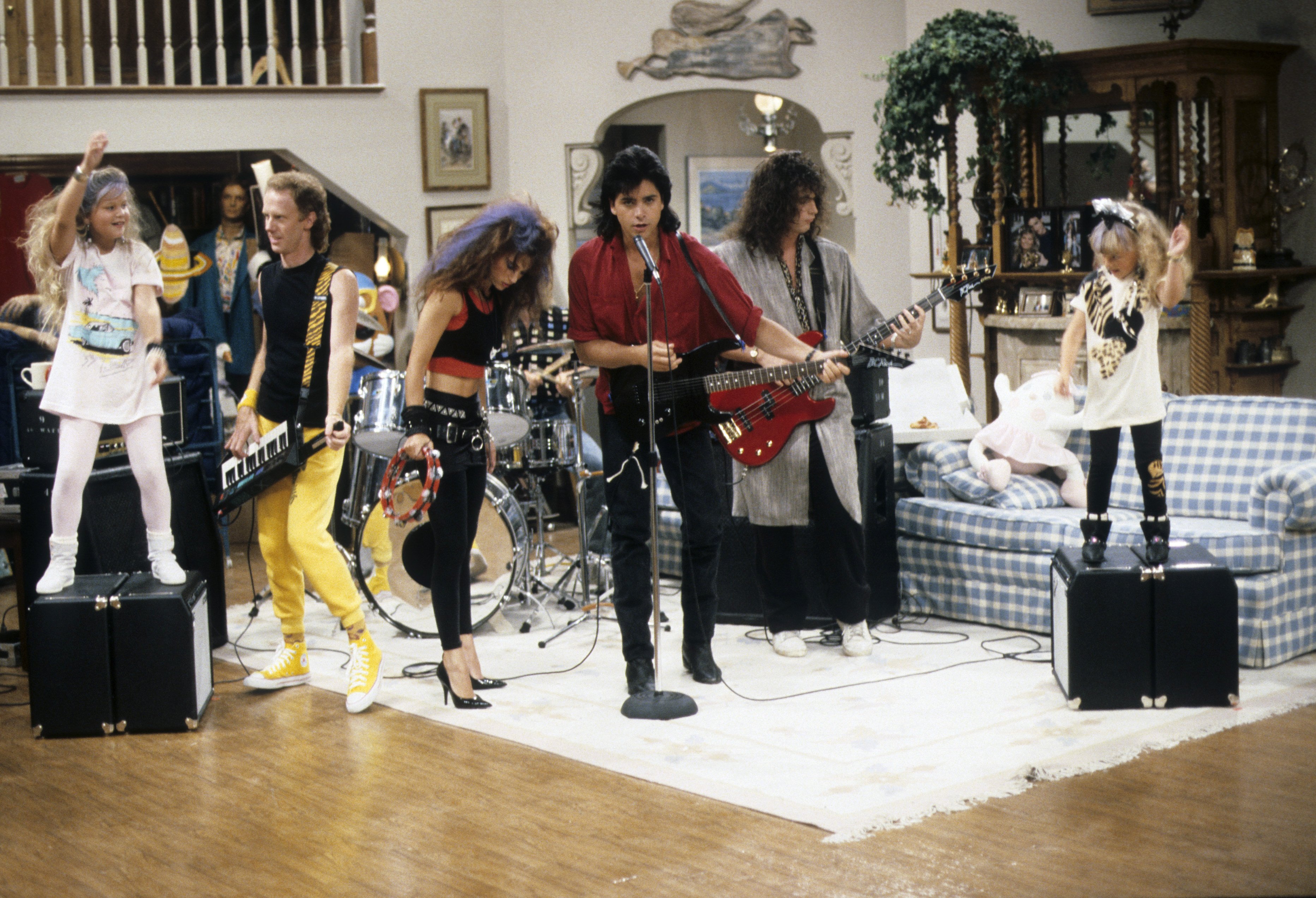 John Stamos commands center stage with an electric guitar as Uncle Jesse rocks out in the Tanner living room, flanked by Candace Cameron Bure, Jodie Sweetin, and his bandmates. Music was central to Stamos' character, reflecting the actor's real-life musical talents as a drummer and performer alongside bands like The Beach Boys and later on Broadway. The energetic scene highlights the charming rock-and-roll spirit he brought to the household, cementing Jesse Katsopolis as a defining figure of '90s television.