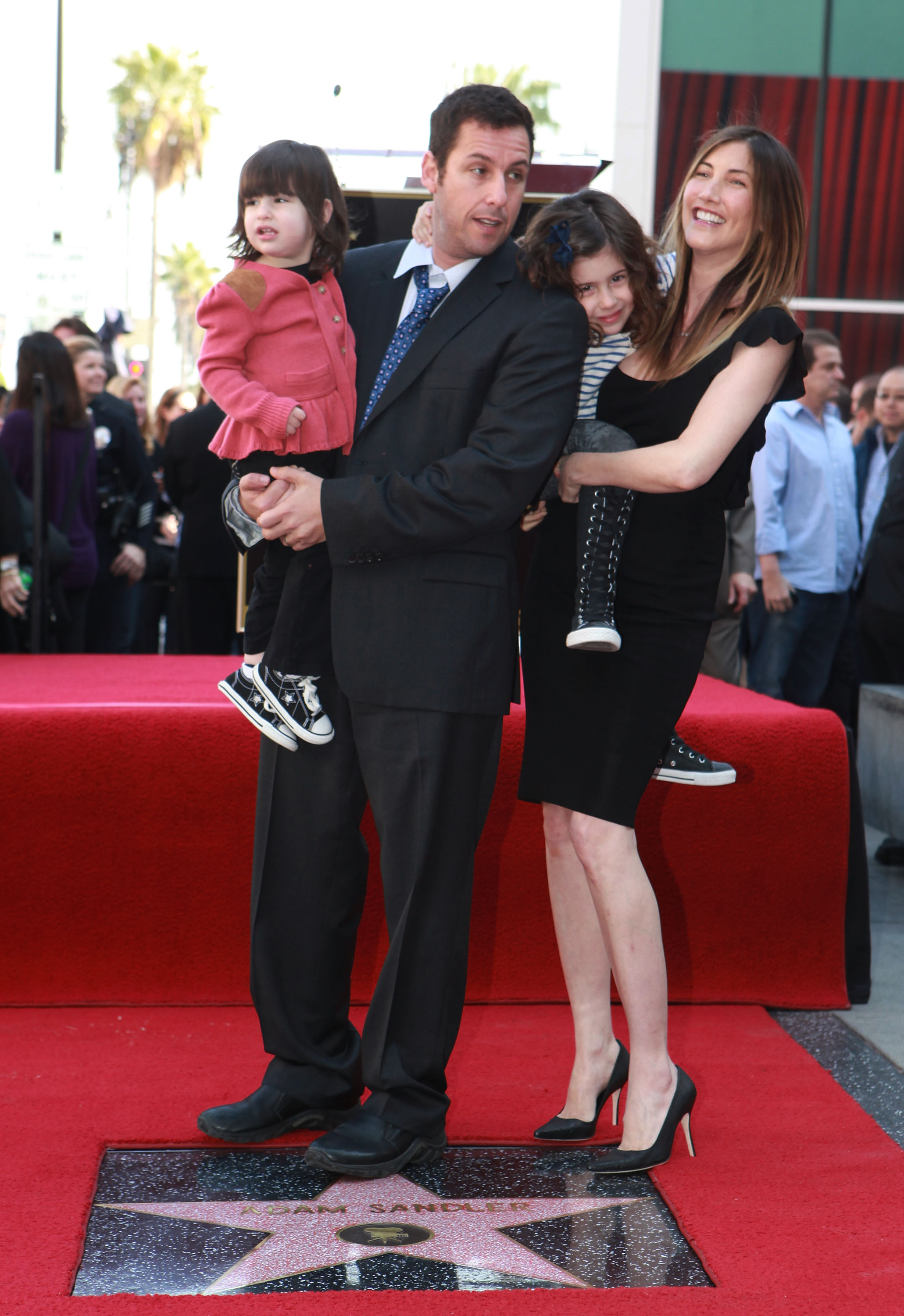 Adam Sandler stands with Jackie and their daughters Sunny and Sadie at his Hollywood Walk of Fame ceremony. Dressed in classic black, Jackie and Adam balance formal elegance with warmth, while the girls add a playful touch — capturing both achievement and family at the heart of it.
