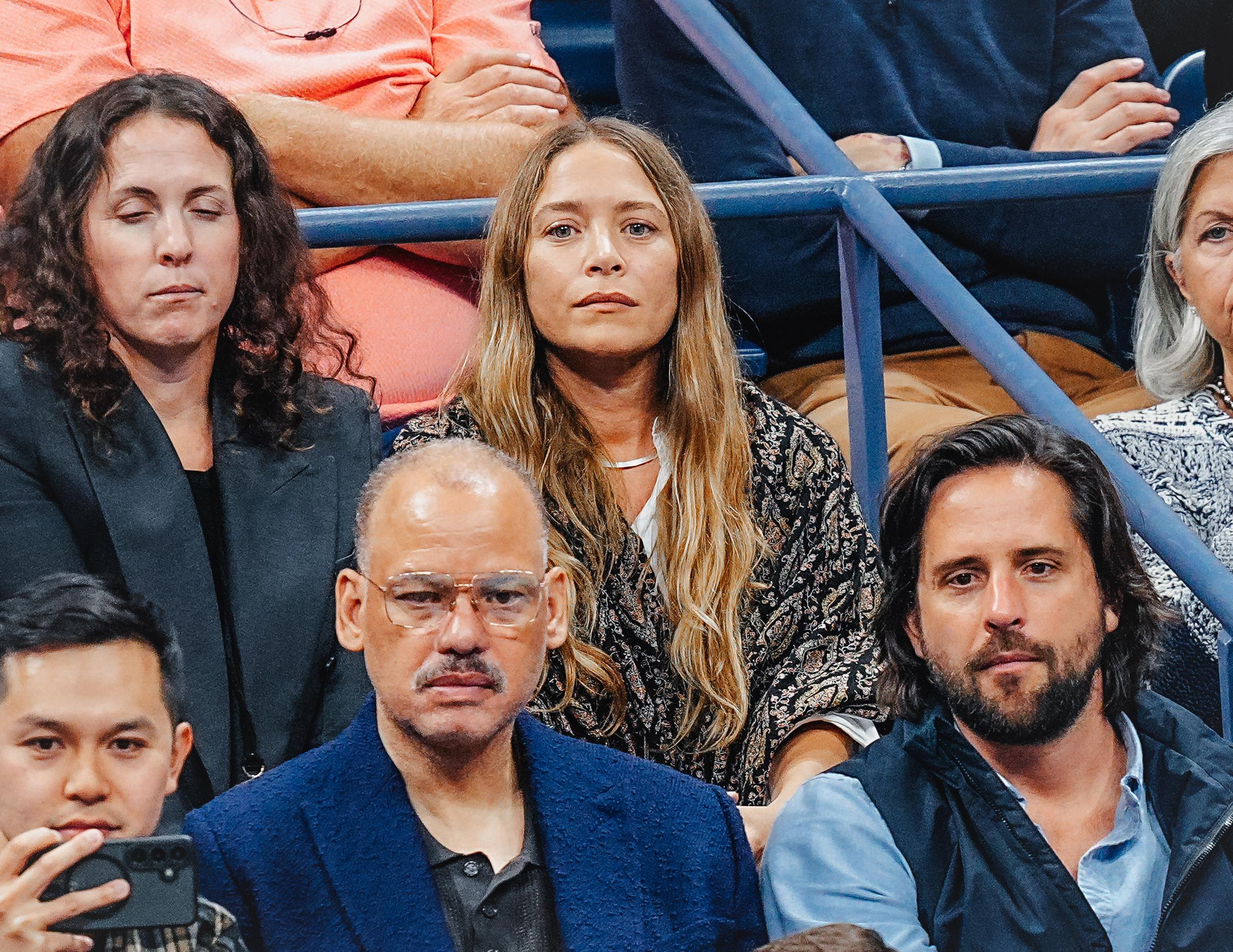Framed by long, flowing wavy blonde-brown hair parted down the center, 39-year-old Mary-Kate Olsen displayed a calm, composed expression with neutral makeup and a subtle silver collar necklace while sitting in the spectator stands at the US Open on September 3, 2025, wearing a dark patterned artisanal jacket over a light top.