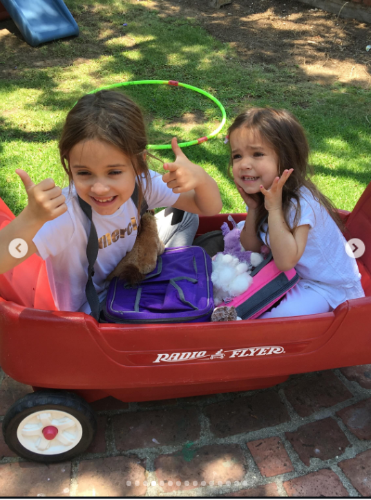 Mabel and Fiona share a playful moment in a large red Radio Flyer wagon, with Mabel giving two thumbs-up while Fiona smiles beside her. A purple bag and several toys sit between them, while a green hula hoop and children’s slide are visible in the sunny backyard behind them. | Source: Instagram/mabellowe_