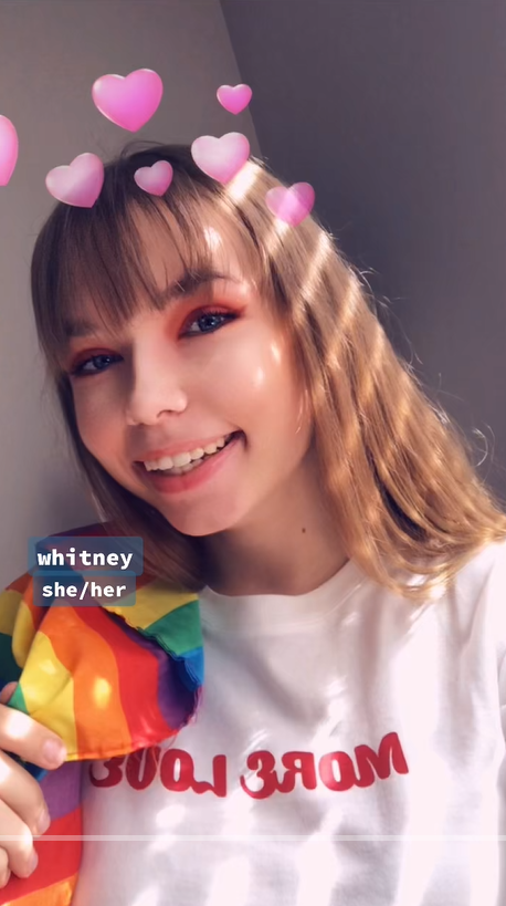 Stevie smiles at the camera with long, light brown hair. She wears a digital flower crown filter, holds a rainbow flag, and is dressed in a white shirt with red lettering. The label on screen identifies her as Whitney with she/her pronouns | Source: TikTok/st0nedc0w