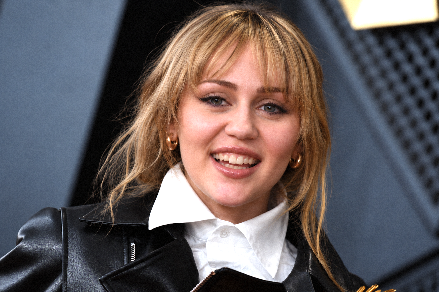 AI-generated image of Miley Cyrus smiling with tousled blonde hair and bangs, wearing a black leather jacket over a crisp white shirt and gold hoop earrings. | Source: Gemini