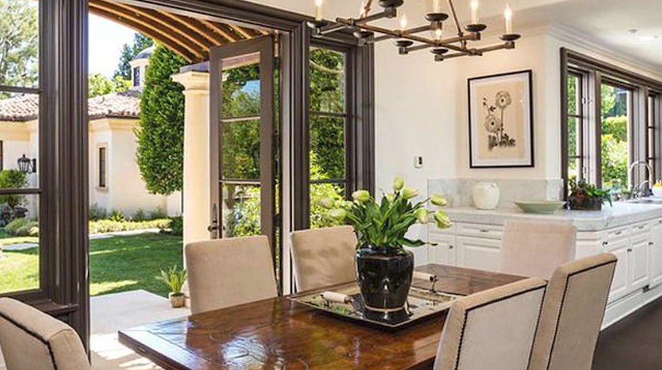 The bright breakfast dining area in Reba McEntire's former Beverly Park home features a polished wood dining table surrounded by upholstered neutral high-back chairs beneath a bronze multi-light chandelier, framed by accordion-style glass doors that fold open directly onto a covered columned veranda and manicured gardens. | Source: YouTube/Taste of Country