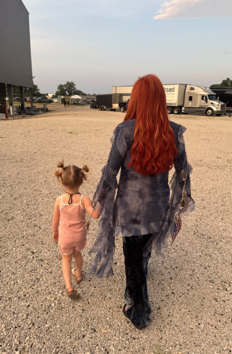 Wynonna holds Kaliyah's hand as they walk across a gravel-covered production area. The little girl wears a pink outfit, while Wynonna's long red hair and flowing stage clothing stand out against the trucks and trailers behind them. | Source: Instagram/wynonnajudd