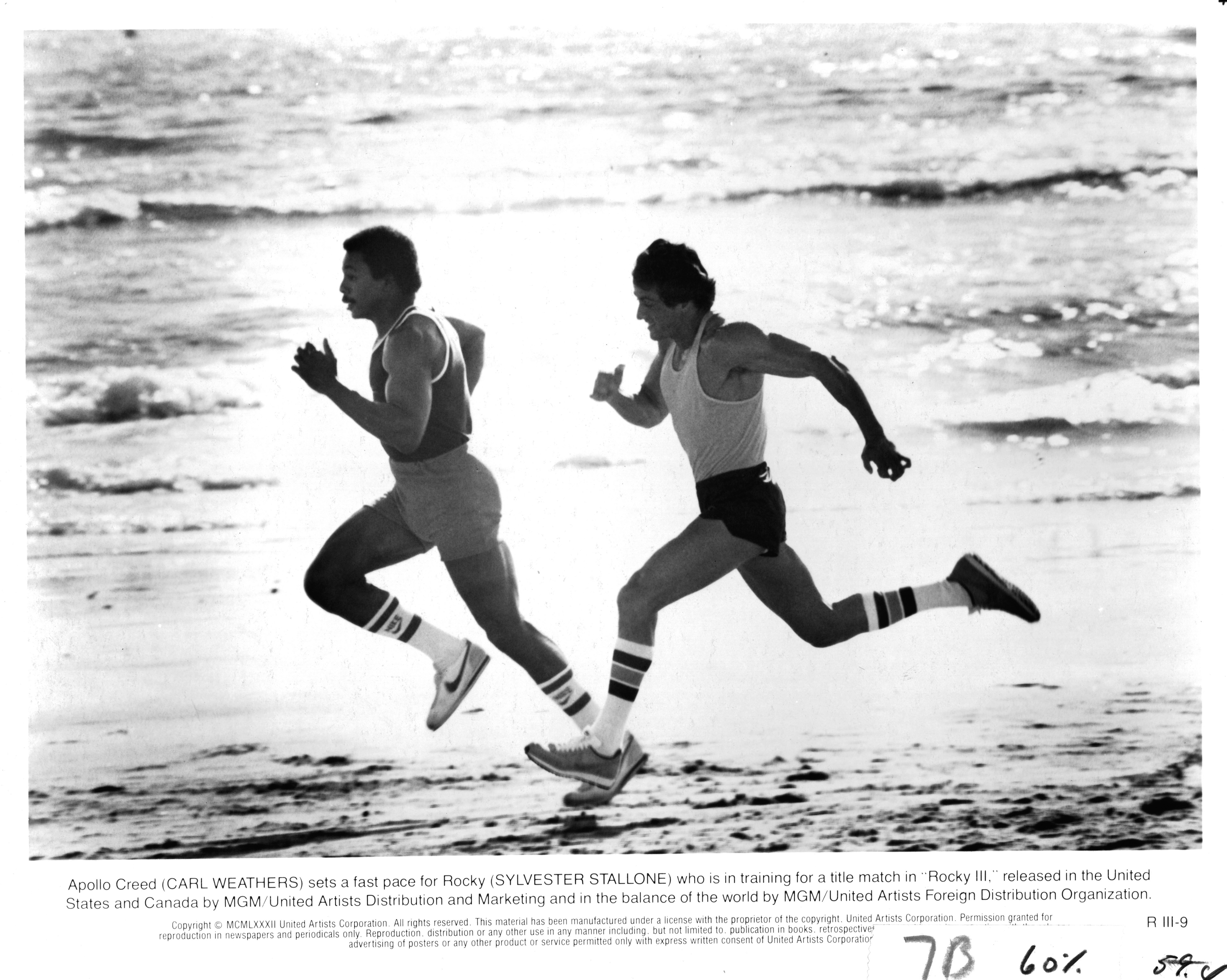 Carl Weathers sets the pace as Apollo Creed while Sylvester Stallone's Rocky Balboa sprints closely behind him along the beach in "Rocky III" in 1982. The training scene highlights Stallone's lean, muscular physique and the greater emphasis on speed and agility as Rocky prepared to face the powerful Clubber Lang.