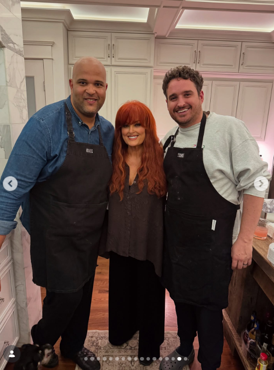Wynonna stands between two men wearing black aprons in a kitchen lined with white cabinets. The photograph captures another food-centered gathering shared through her social media account. | Source: Instagram/wynonnajudd