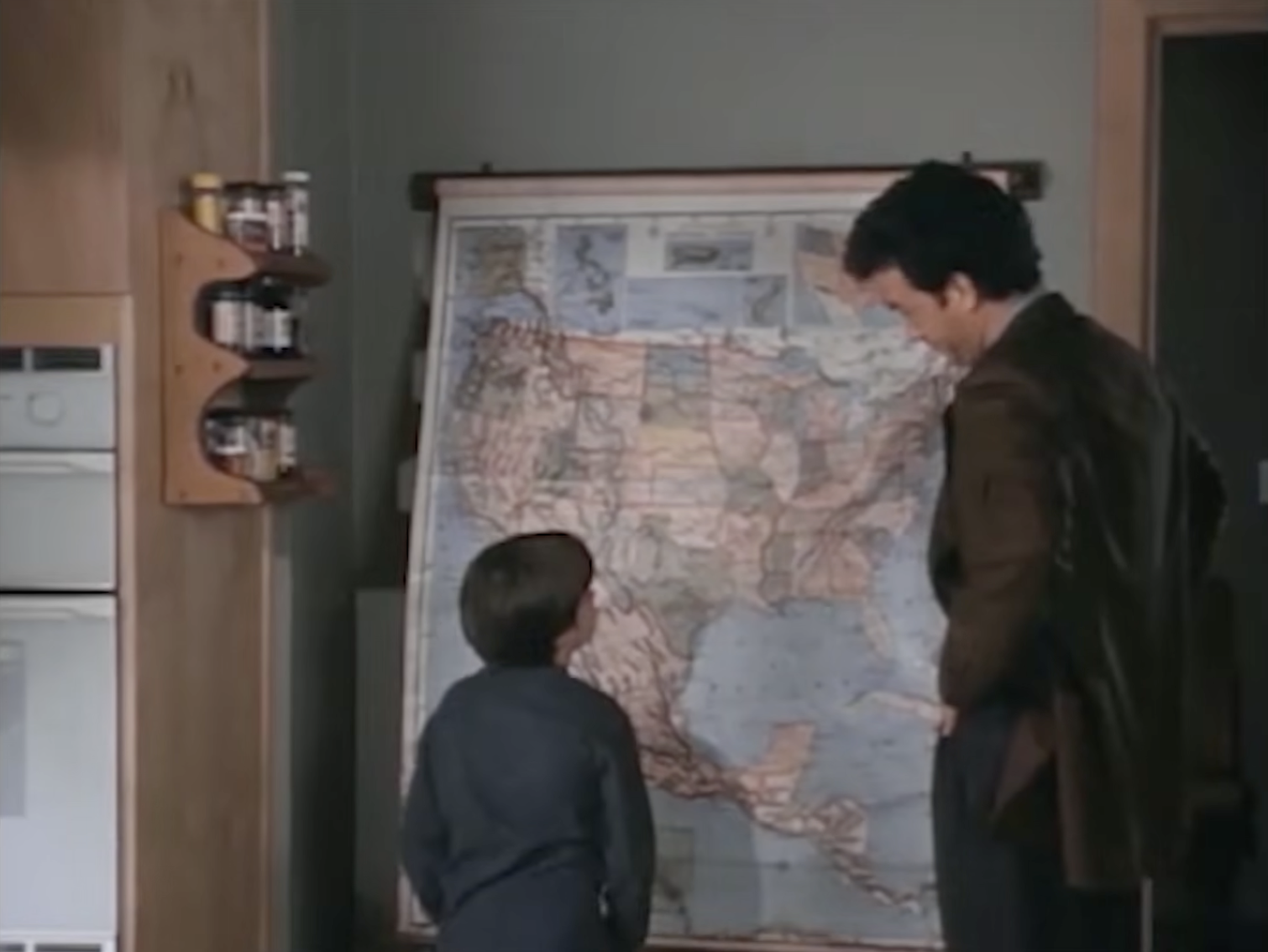 A young boy studies a map of the United States with his on-screen father in a scene from Sleepless in Seattle, capturing a quiet moment between the two characters in the beloved romantic comedy. | Source: Rotten Tomatoes Classic Trailers / YouTube.