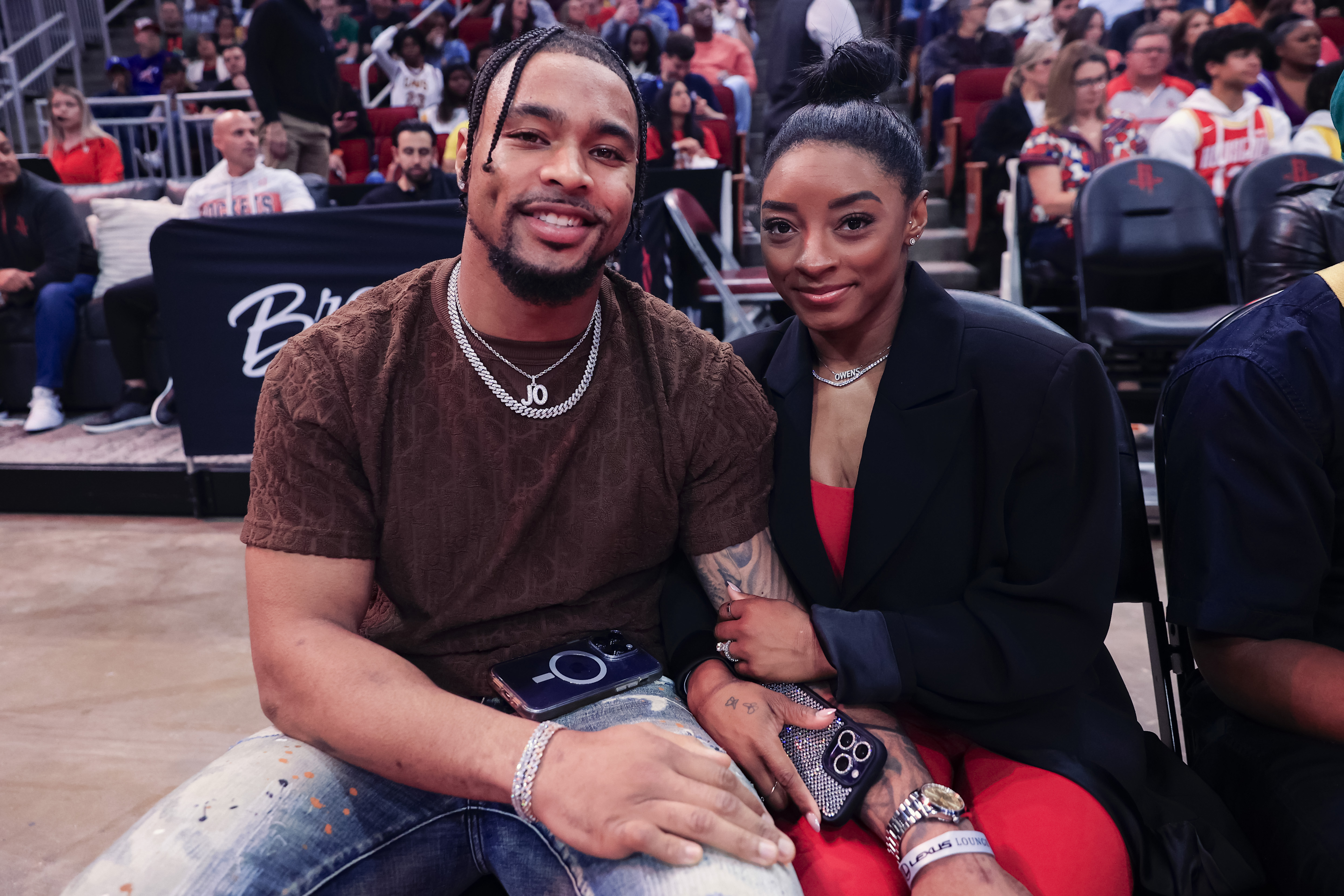 Simone Biles and Jonathan Owens sit courtside during a game between the Houston Rockets and the Los Angeles Lakers at the Toyota Center in Houston, Texas, on January 29, 2024. Biles sports a red outfit layered under a black blazer, while Owens wears a brown patterned shirt and distressed jeans.