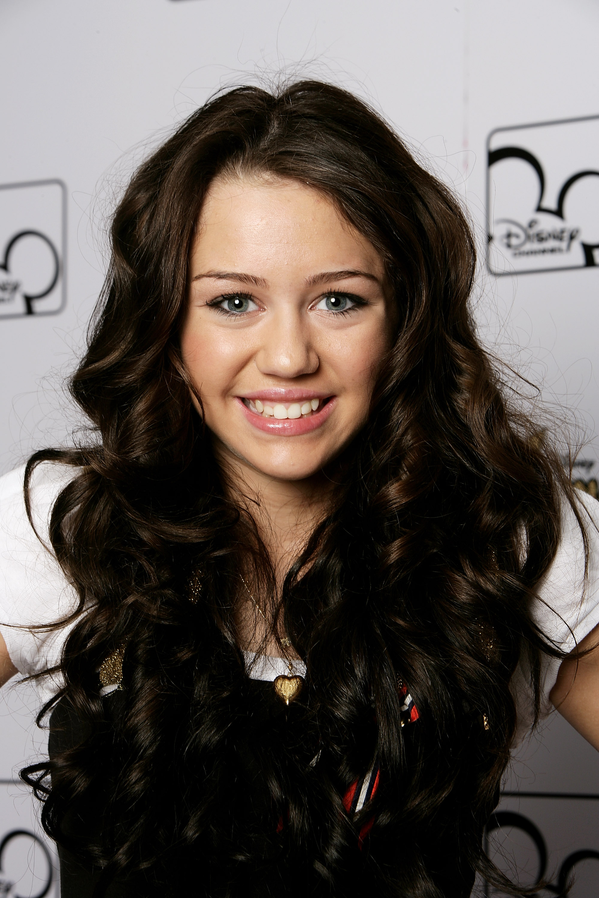 Miley Cyrus posed in front of a Disney Channel-branded backdrop with long, voluminous dark curls, a white top, and a gold heart pendant. The portrait reflects the polished, youthful image associated with her rise to fame as a Disney star. | Source: Getty Images