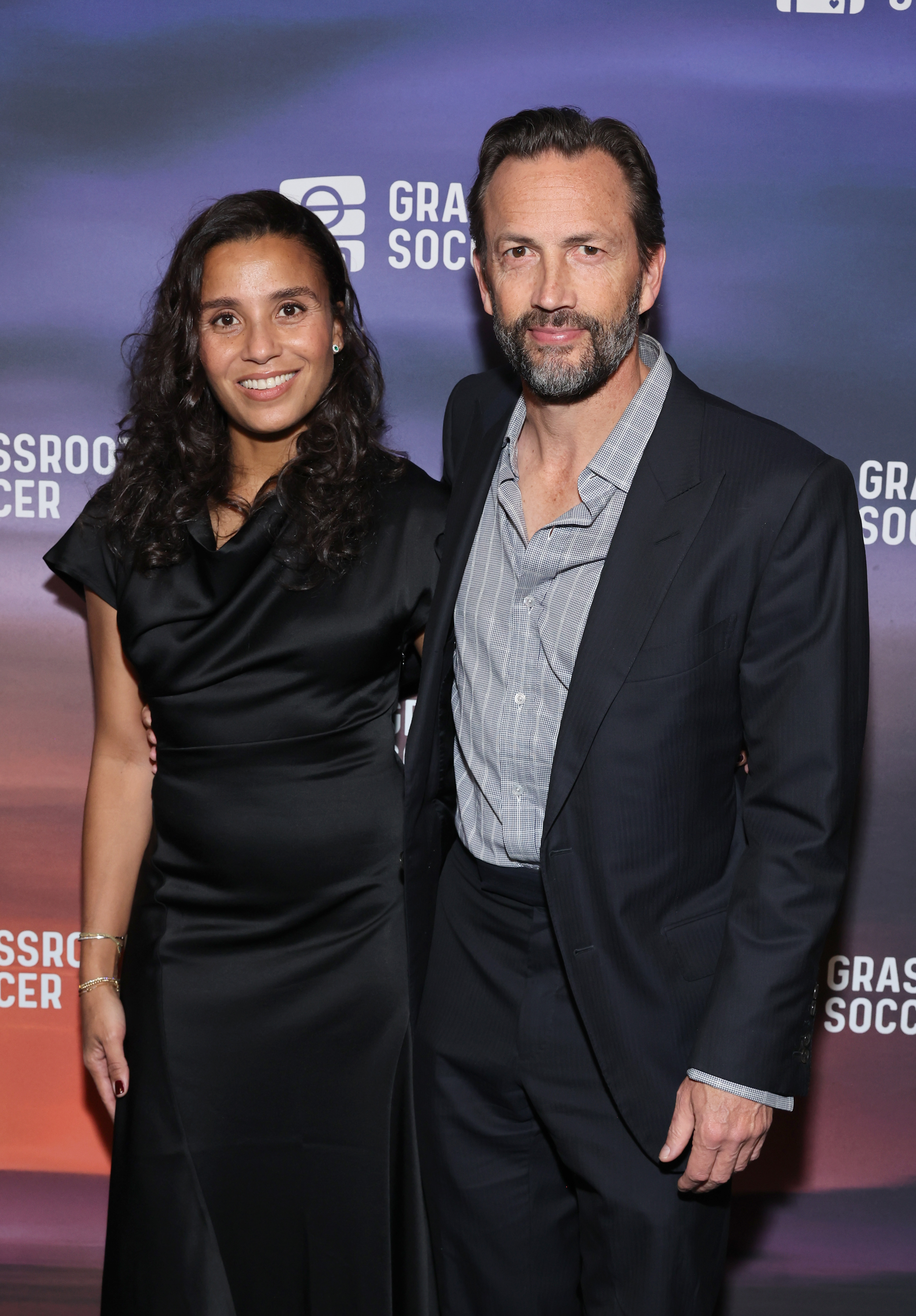Marilee Fiebig and Andrew Shue attend the 2024 Grassroot Soccer World AIDS Day Gala at Hall des Lumières in New York City on December 3, 2024. The couple smiles together on the red carpet, with Fiebig wearing a chic black cowl-neck satin dress accented with gold jewelry, while Shue coordinates beside her in a dark navy tailored suit layered over a textured open-collar button-down shirt. | Source: Getty Images