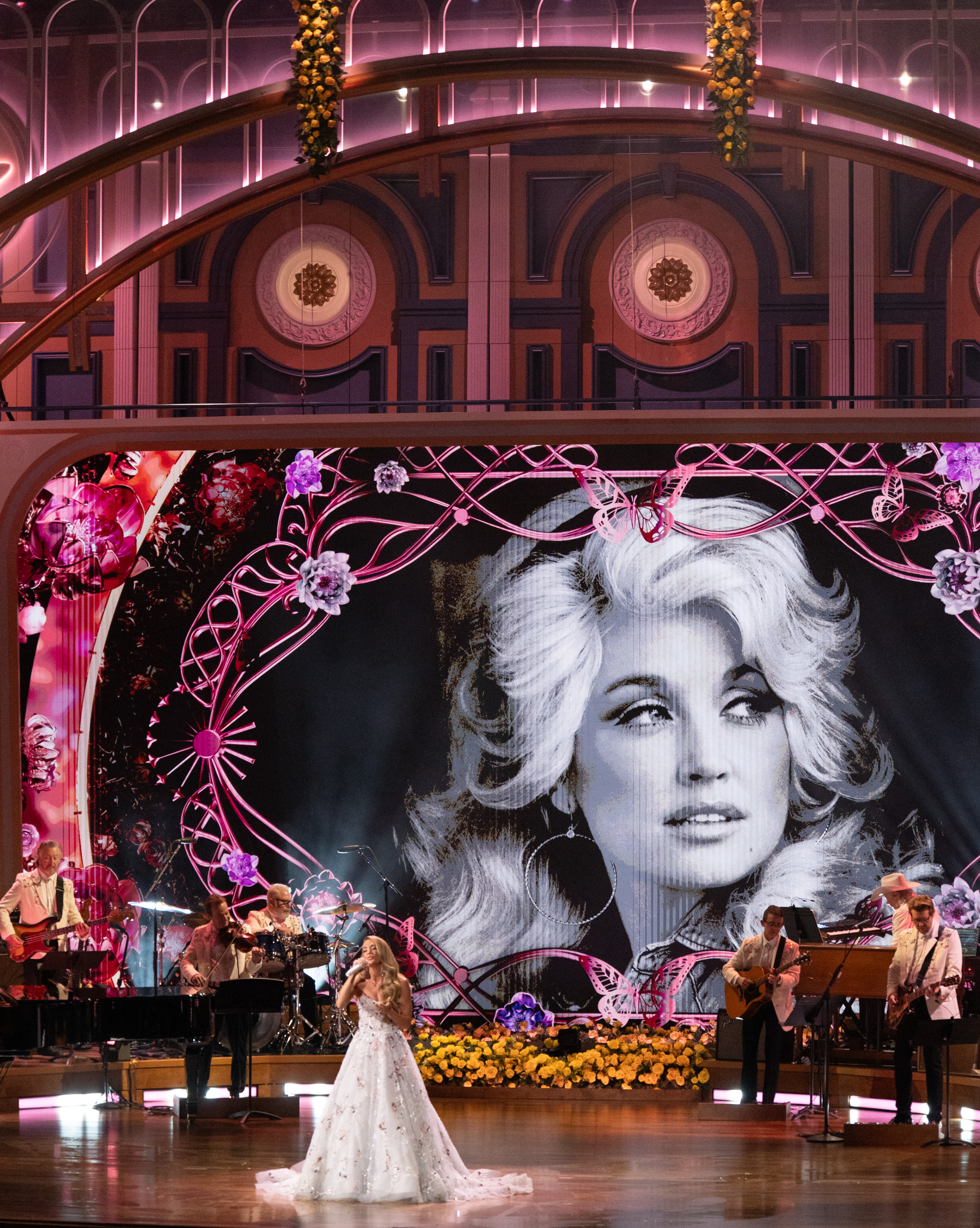 Carrie Underwood performs beneath a giant black-and-white portrait of Dolly Parton, surrounded by pink butterflies and floral decorations during the Nashville tribute. | Source: Getty Images.