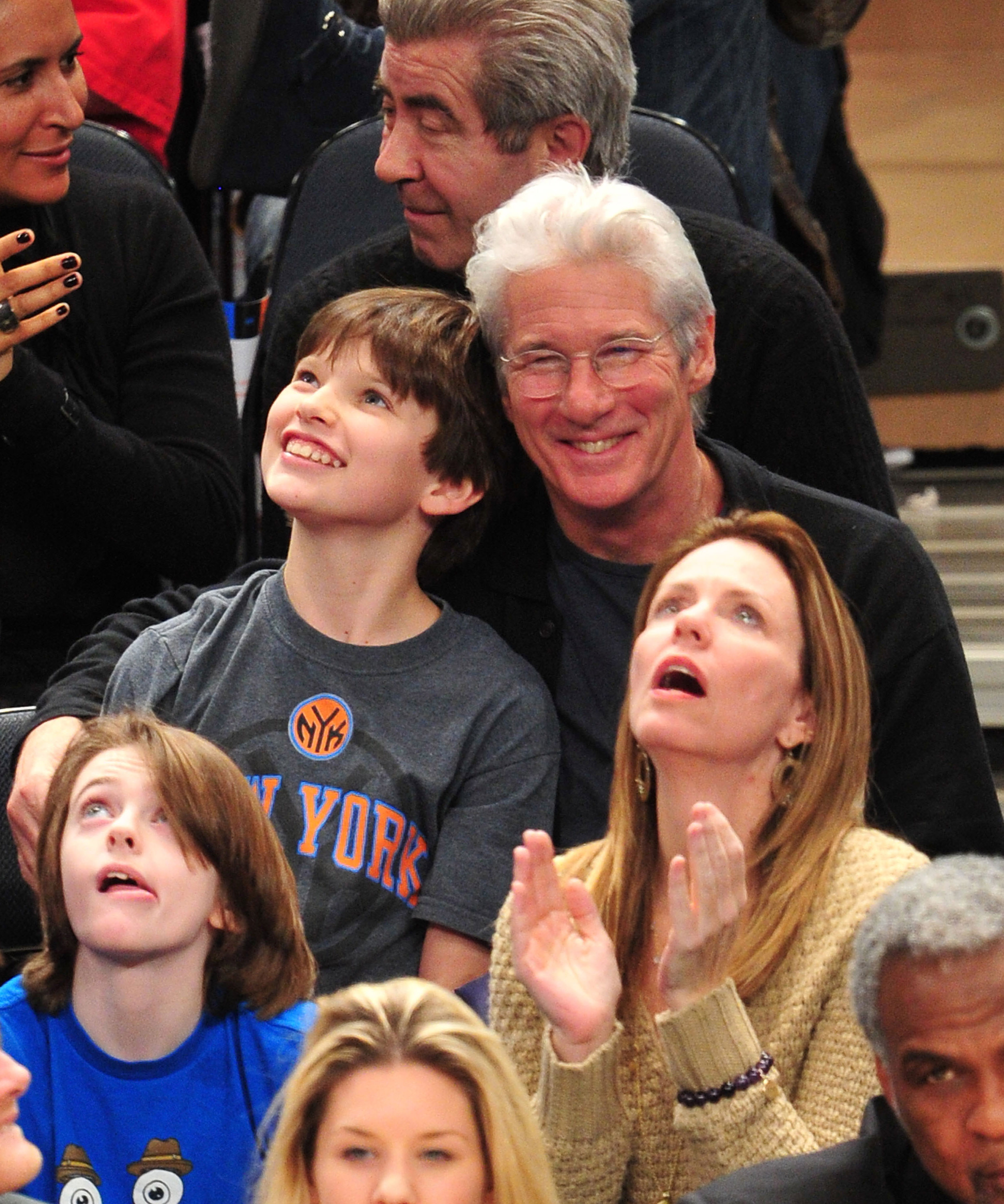 Beaming with joy, Richard Gere leans in close to Homer Gere during a Knicks game at Madison Square Garden in New York City, sharing a candid courtside moment surrounded by fellow fans.