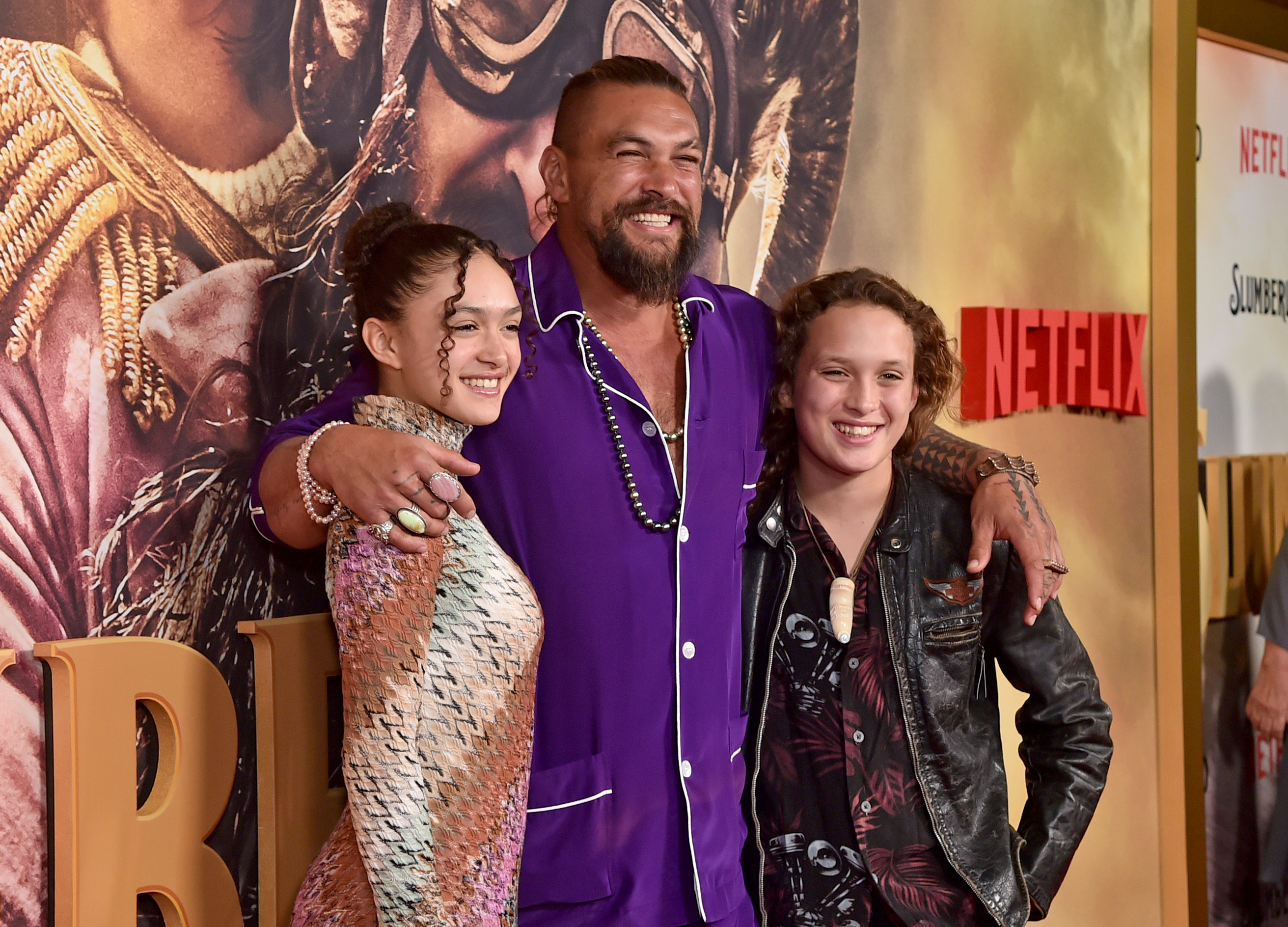 Jason Momoa shares a cheerful moment with daughter Lola Iolani Momoa and son Nakoa-Wolf Manakauapo Namakaeha Momoa at the Los Angeles premiere of Netflix's "Slumberland" at AMC Century City 15 on November 9, 2022, in Century City, California. Momoa sports a buzz cut with a fade, beaded necklaces, and a vibrant royal-purple silk pajama-style shirt with white piping, wrapping his arms around Lola in a textured knit chevron-patterned turtleneck dress and Nakoa-Wolf in a rugged leather jacket over a floral shirt with a carved pendant necklace. | Source: Getty Images