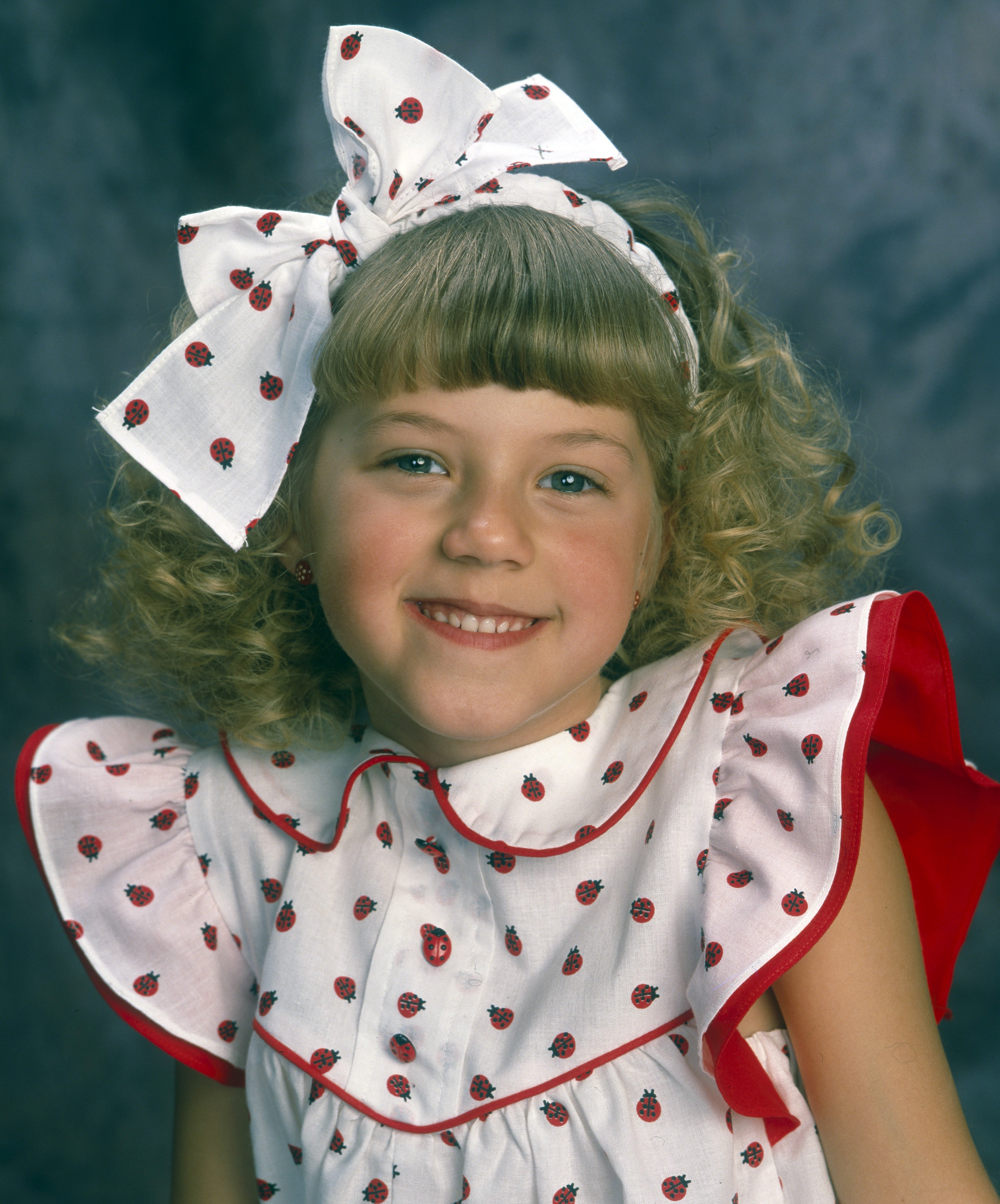 Jodie Sweetin smiles warmly in a promotional gallery portrait for "Full House" from March 1, 1989, wearing a ladybug-print dress paired with an oversized matching hair bow. Bringing endless personality, comic timing, and sweetness to middle sister Stephanie Tanner, Sweetin became an immediate fan favorite whose memorable catchphrases and childhood adventures endeared her to millions of viewers throughout the sitcom's original run.