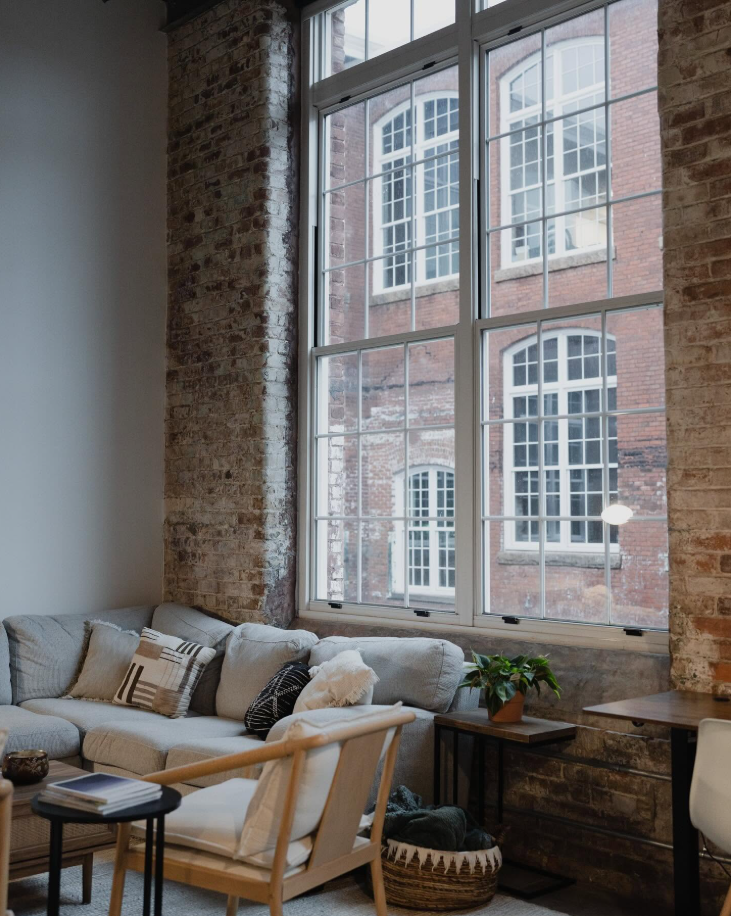 A cozy living area inside a Judson Mill Lofts apartment features a plush sectional, exposed brick walls and oversized industrial-style windows overlooking the historic mill buildings. | Source: Facebook/Judson Mills Loft