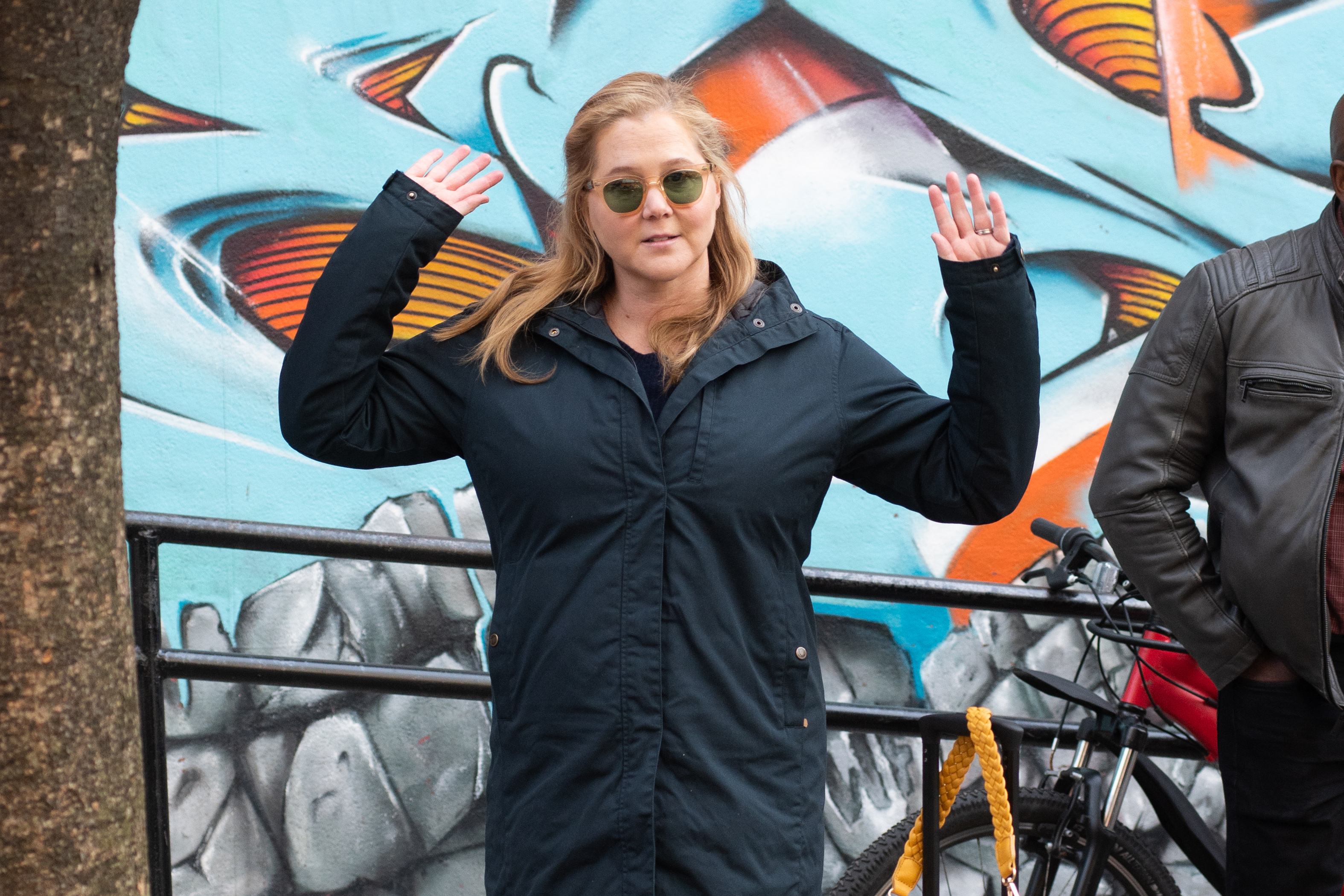 Amy Schumer raises her hands while filming a scene for "Life & Beth" in New York City. She wears a navy jacket and sunglasses, standing in front of a colorful mural.