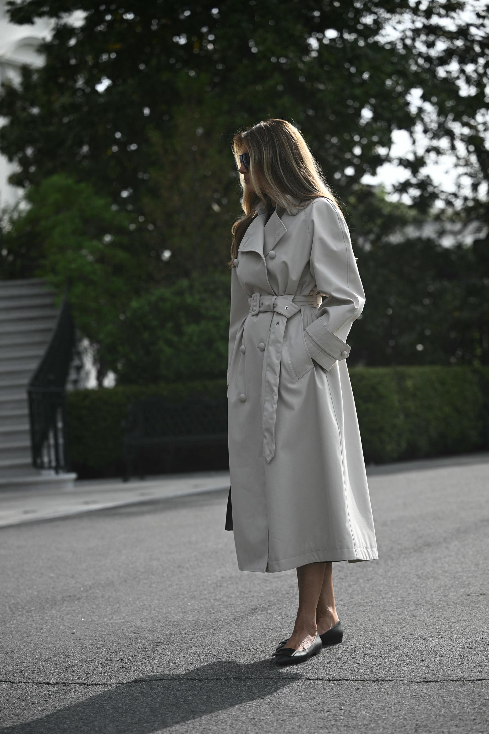First Lady Melania Trump stands on the South Lawn of the White House in Washington, D.C., on April 25, 2025, departing for Joint Base Andrews en route to Rome, wearing a belted, double-breasted stone trench coat and dark sunglasses, paired with understated black leather pointed-toe ballet flats that bring functional polish to her travel attire.