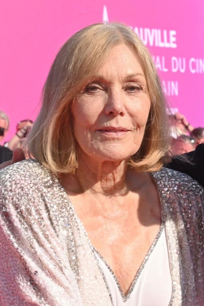 Kim Novak poses with both hands resting near her chest, wearing a white V-neck dress and a shimmering silver jacket as a pink event backdrop frames the portrait. | Source: Gemini