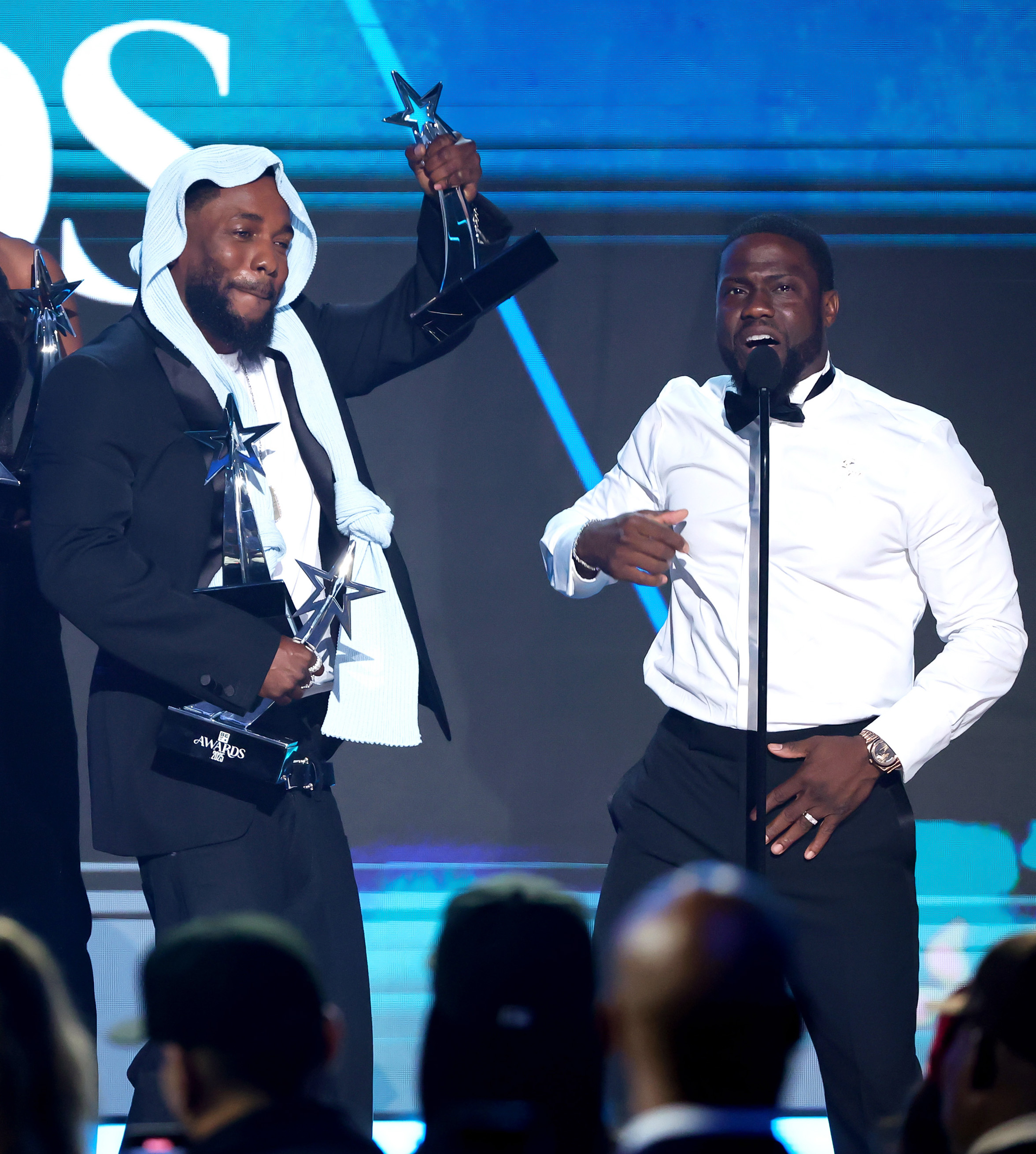 Holding up one of his trophies, Kendrick Lamar stands next to Kevin Hart as they share a lighthearted moment at the 2025 BET Awards. Lamar's multiple wins became a running joke as Hart attempted to help him carry the load.