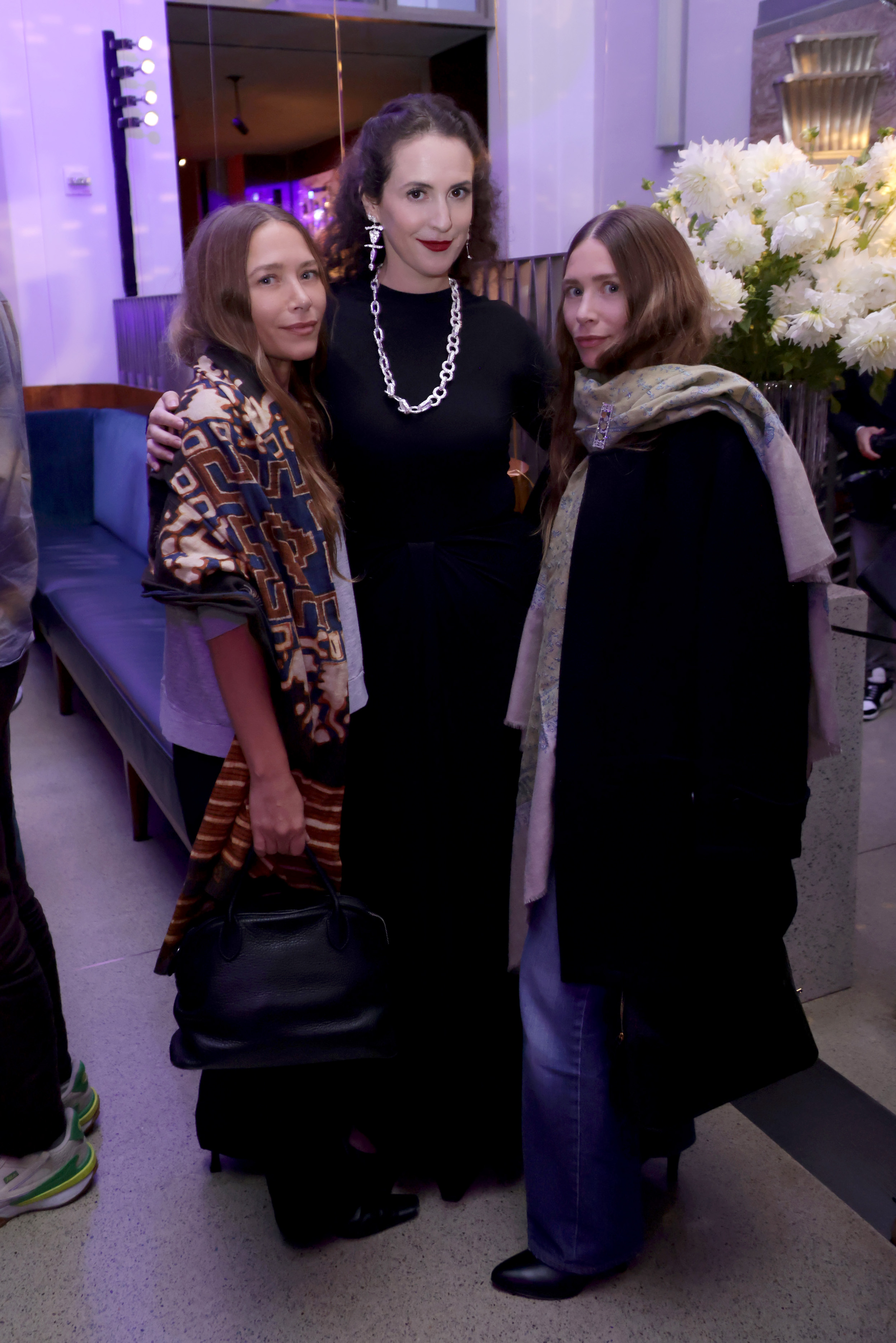 Natural smiles and relaxed facial expressions accompanied 39-year-old Mary-Kate and Ashley Olsen alongside Sara Moonves at a New York Fashion Week celebration at Eleven Madison Park Restaurant on September 11, 2025, where Mary-Kate wore a patterned scarf over a dark outfit with a black handbag, and Ashley sported loose blonde-brown locks draped with a patterned scarf, a dark coat, and blue jeans.