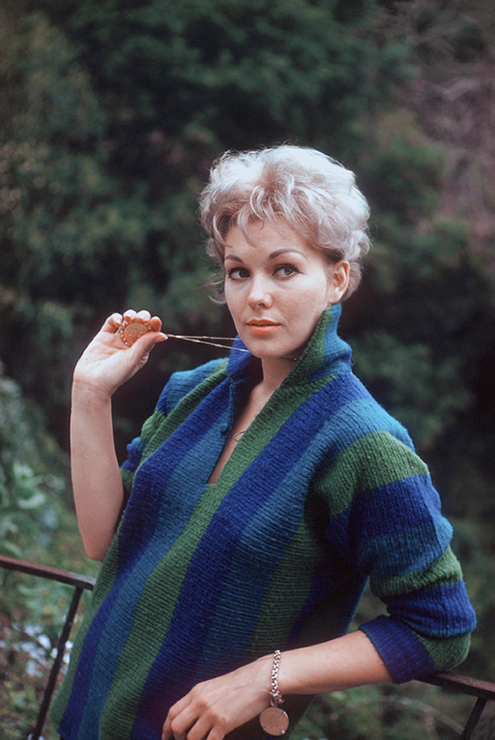 Kim Novak poses at home in a colorful blue-and-green knitted outfit, holding a pendant near her face. | Source: Getty Images