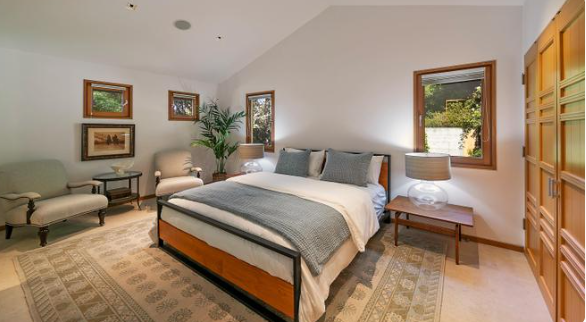 The bedroom features a large bed with gray and white bedding, wooden furniture, built-in wardrobes, and a cozy seating area beside windows overlooking the greenery. | Source: Redfin