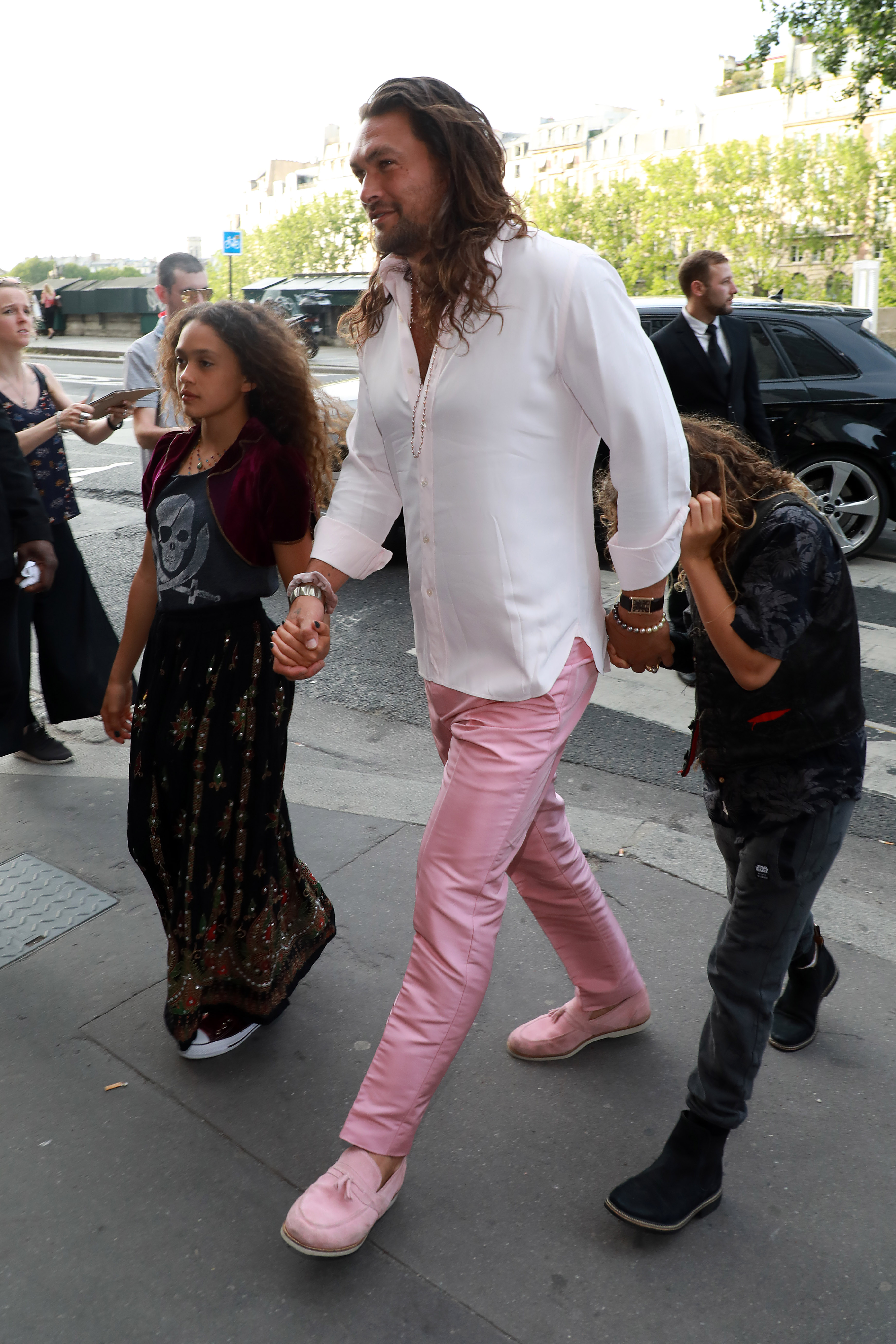 Jason Momoa holds hands with daughter Lola Iolani Momoa and son Nakoa-Wolf Manakauapo Namakaeha Momoa as they arrive at Lapérouse restaurant for Zoë Kravitz and Karl Glusman's wedding celebration dinner on June 28, 2019, in Paris, France. Momoa strides through the street in a crisp white button-down shirt paired with pastel pink trousers and matching pink suede loafers, walking alongside Lola in an embroidered maxi skirt with a skull-print top and velvet bolero, while Nakoa-Wolf follows closely in dark casual layers. | Source: Getty Images