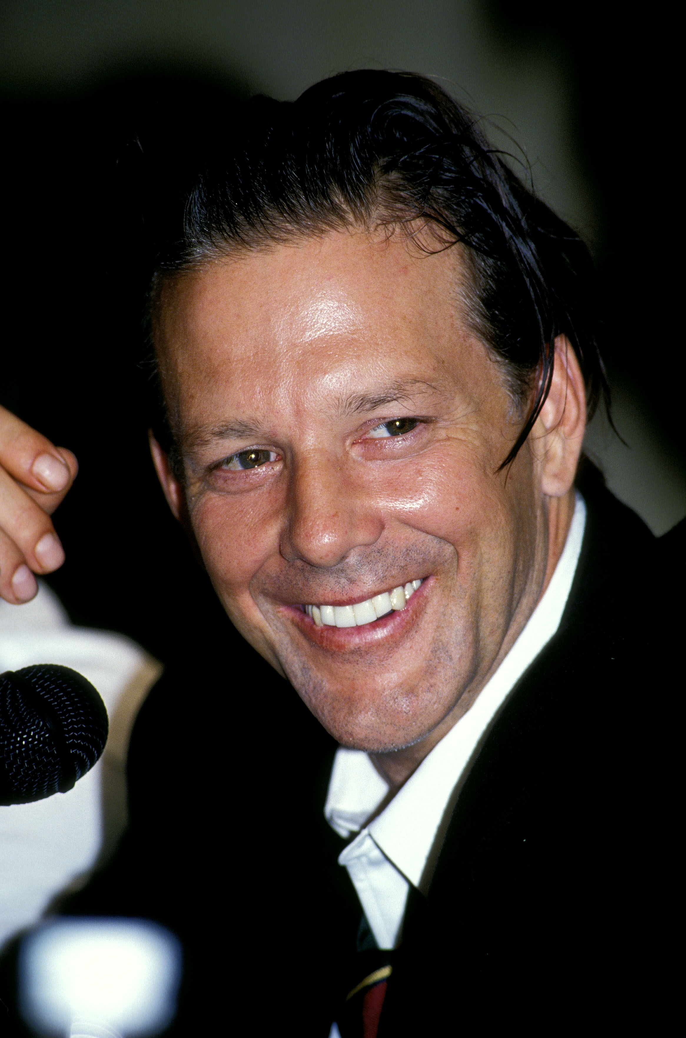 Mickey Rourke beams with a charismatic smile during a press conference in Buenos Aires. His slicked-back hair and sharp suit reflect both charm and confidence as he engages with the crowd.