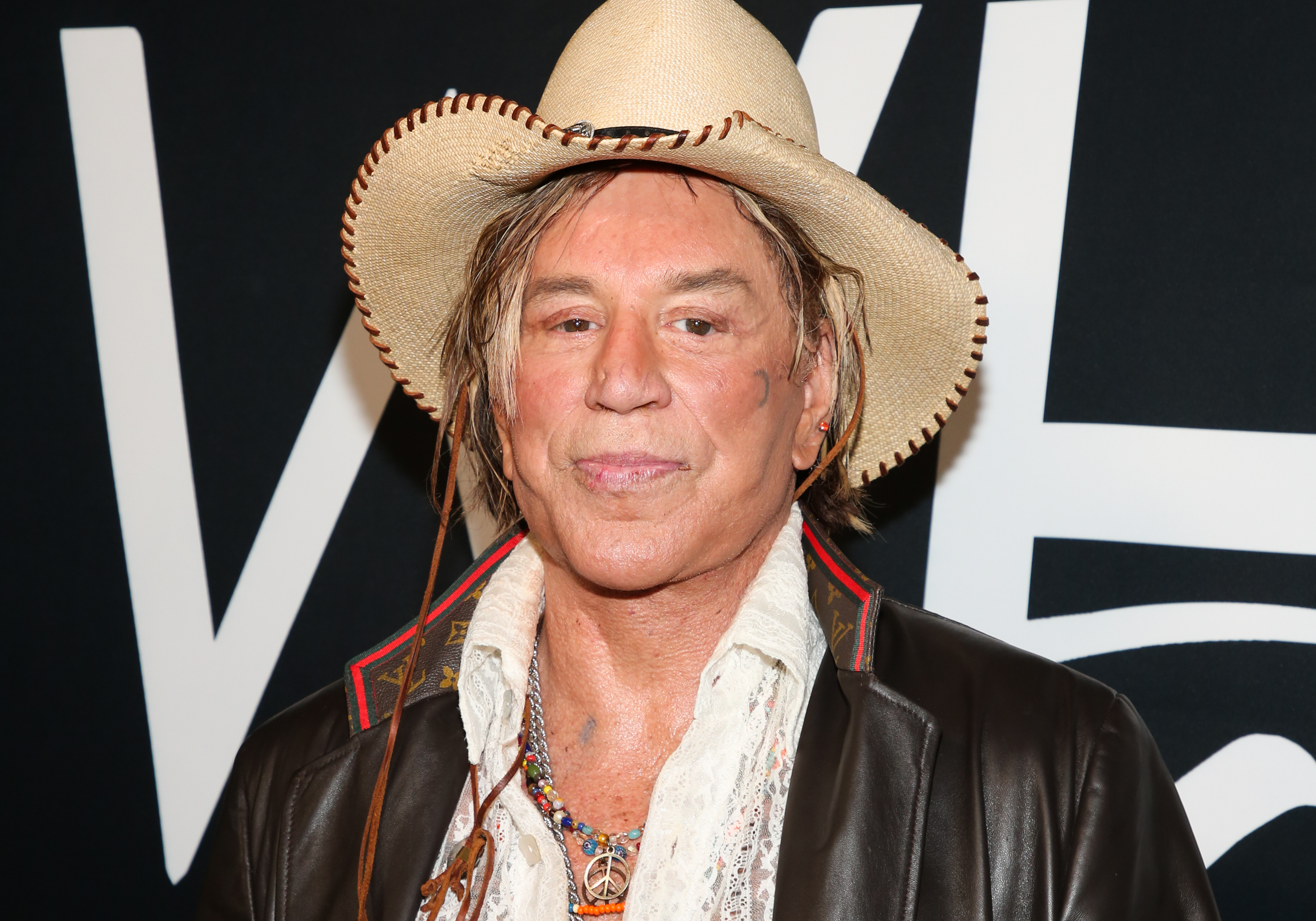 Mickey Rourke attends the TEATRO Even Summer White Affair at Skybar in West Hollywood, California, wearing a wide-brimmed cowboy hat, layered necklaces, and a leather jacket with flair. His eclectic style and steady gaze command attention, blending rugged charm with artistic eccentricity.