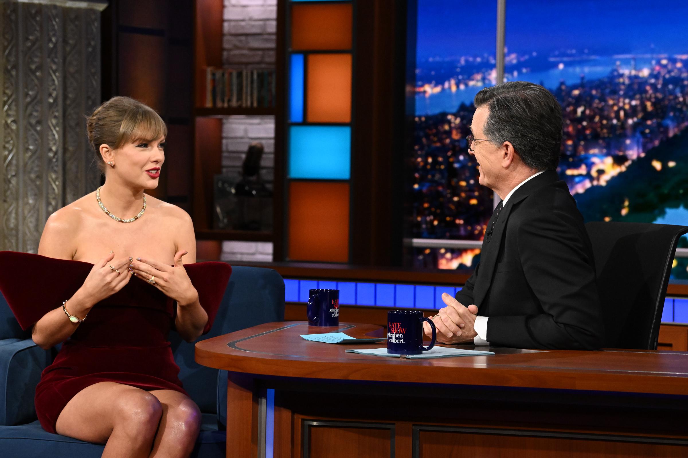 Seated opposite Stephen Colbert, Taylor Swift gestures expressively during their interview, clad in her structured David Koma velvet minidress. The conversation covered her music and recent tour milestones, set against the show's colorful set.