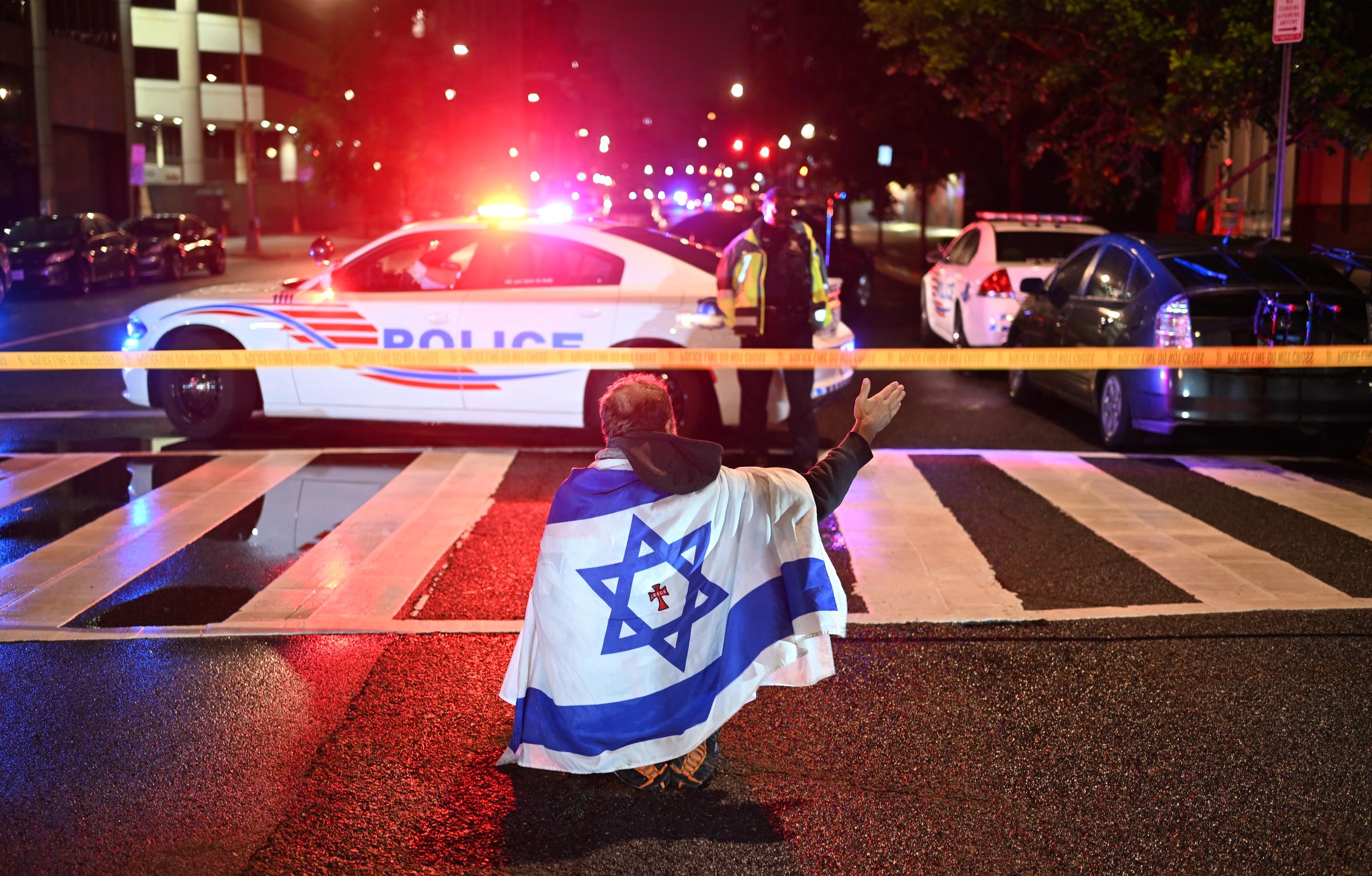 A man draped in the Israeli flag, bearing a cross and the name "Jesus" at its center, gestures as Metropolitan Police officers secure the area outside the Capital Jewish Museum, in Washington D.C., on May 22, 2025 | Source: Getty Images
