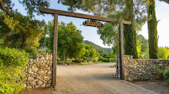 A rustic wooden arch bearing the name "Indian Way" marks the entrance to the property, with stone walls, a gravel driveway, and trees surrounding the gate. | Source: Redfin