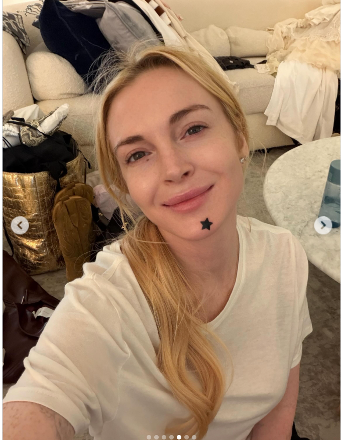 Lindsay Lohan poses for a makeup-free selfie indoors, wearing a simple white T-shirt with her blonde hair swept over one shoulder. A small black star is visible on her chin as she smiles at the camera. | Source: Instagram/lindsaylohan
