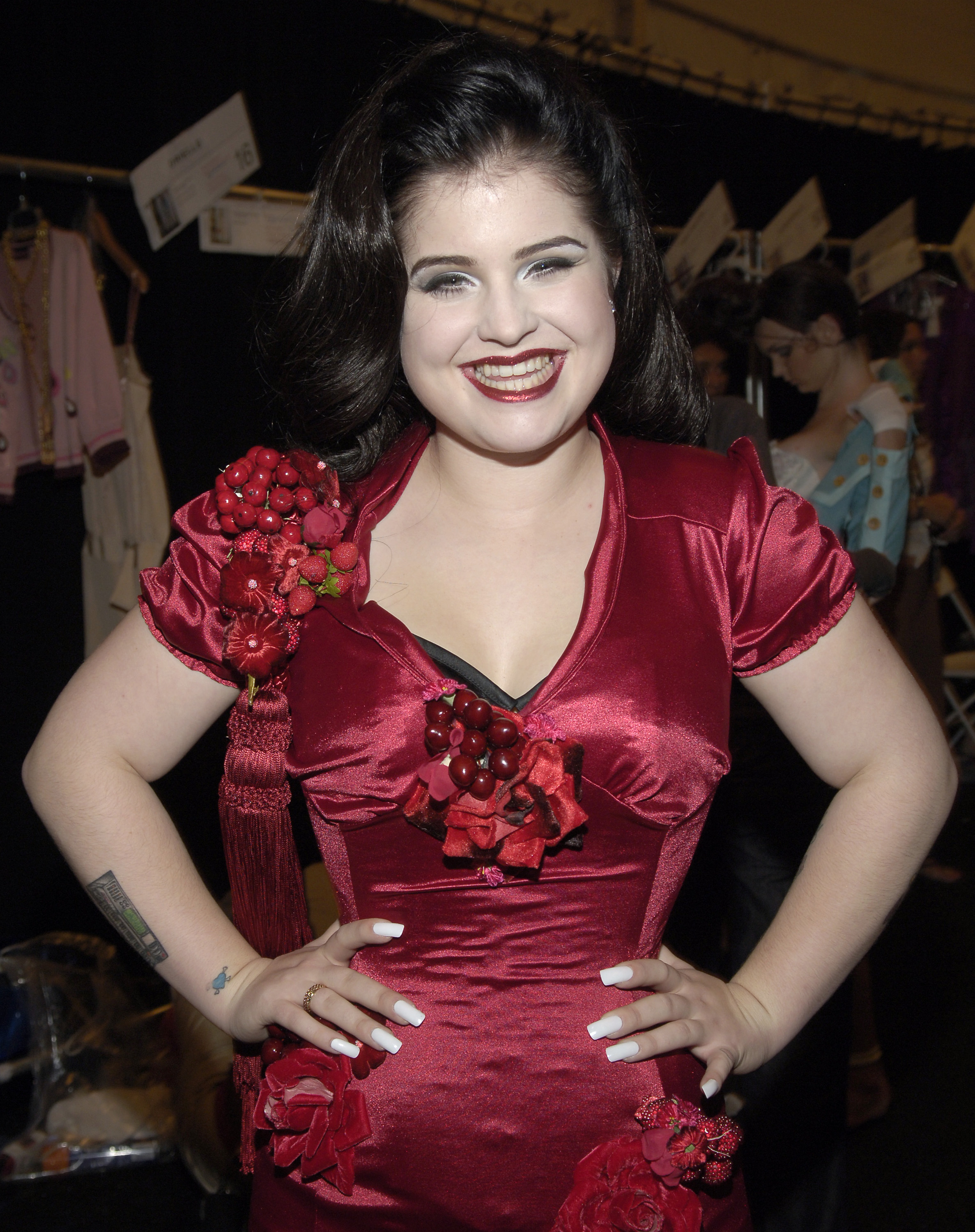 Kelly Osbourne poses backstage at the Heatherette Spring 2006 fashion show during Olympus Fashion Week in New York City, embracing retro glamour with voluminous dark hair, bold makeup, and a striking crimson dress adorned with floral details. | Source: Getty Images